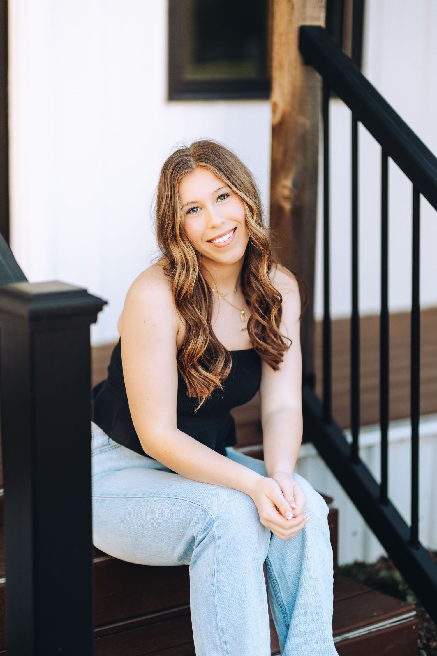 Smiling young woman with wavy auburn hair sitting on porch steps in black top and jeans.