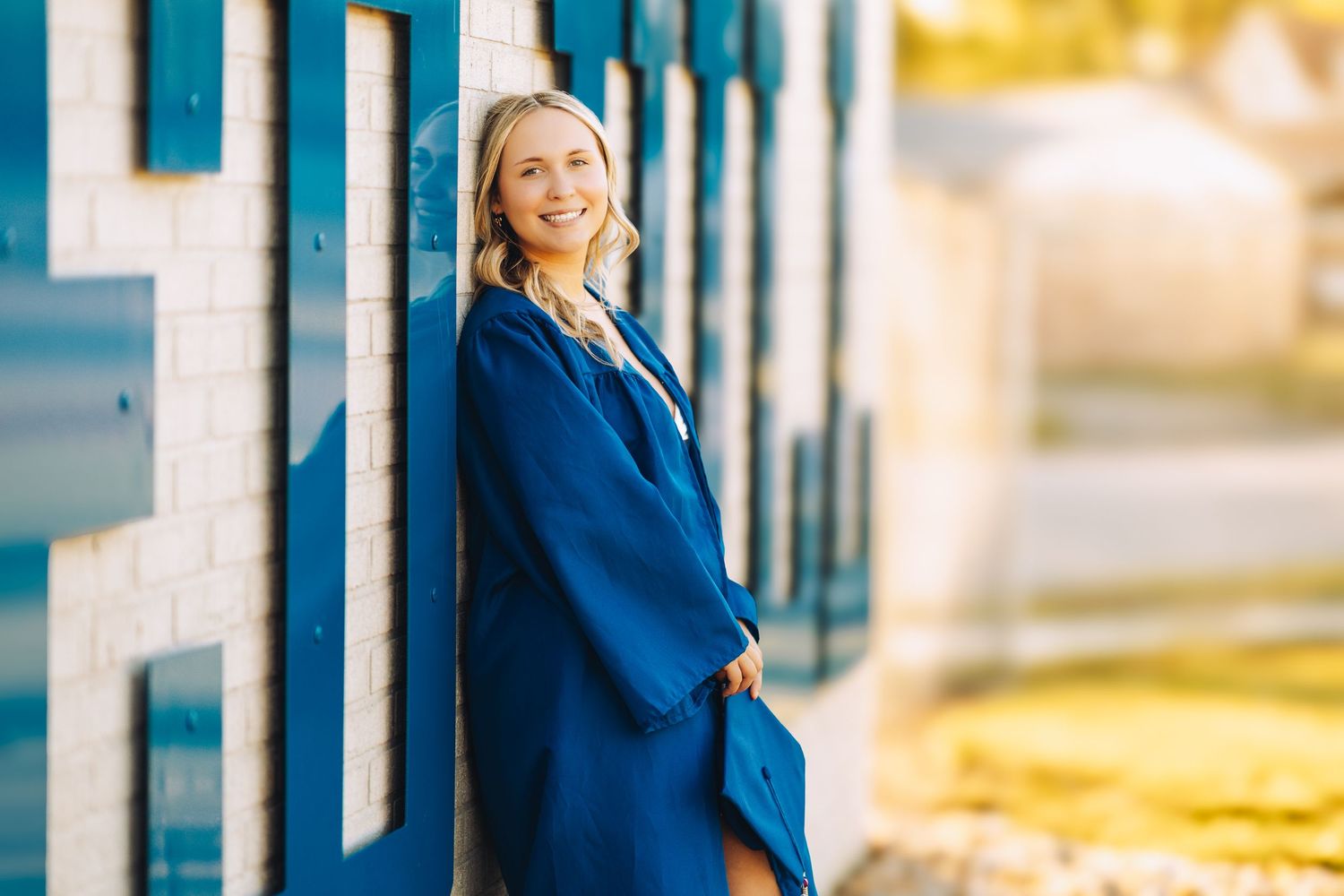 Smiling blonde woman in blue coat leaning against a blue and white tiled wall outdoors.