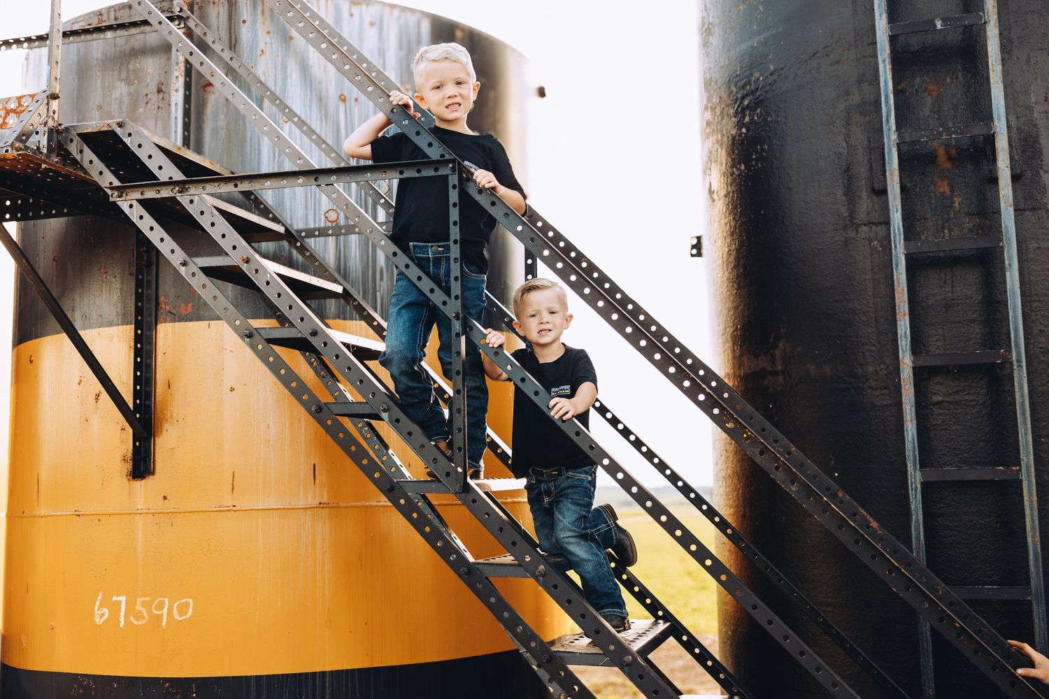 Two young boys in matching outfits climb a metal industrial staircase on a yellow grain silo.