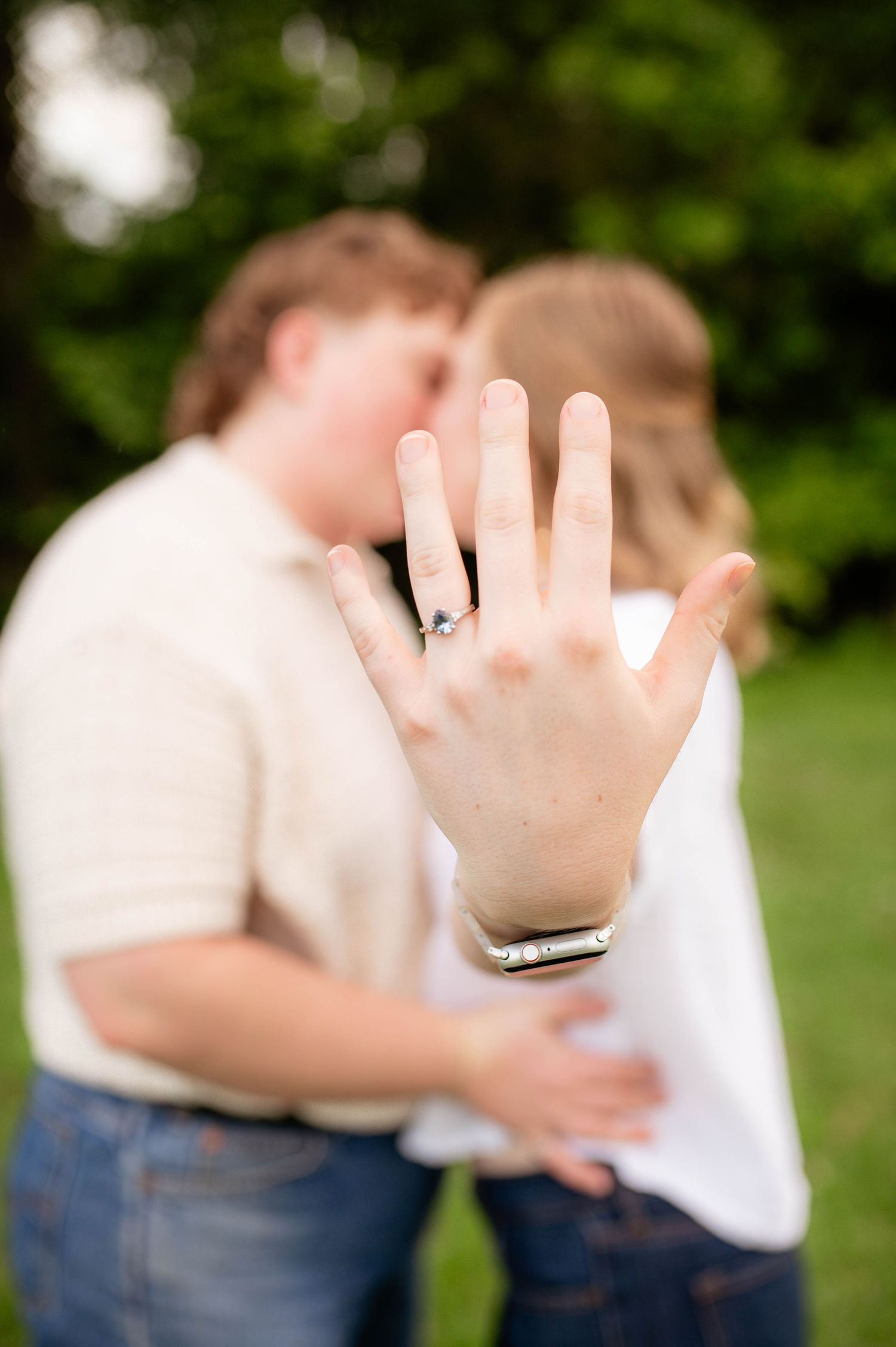 Two engaged people, one showing off her ring while her partner snuggles into her.