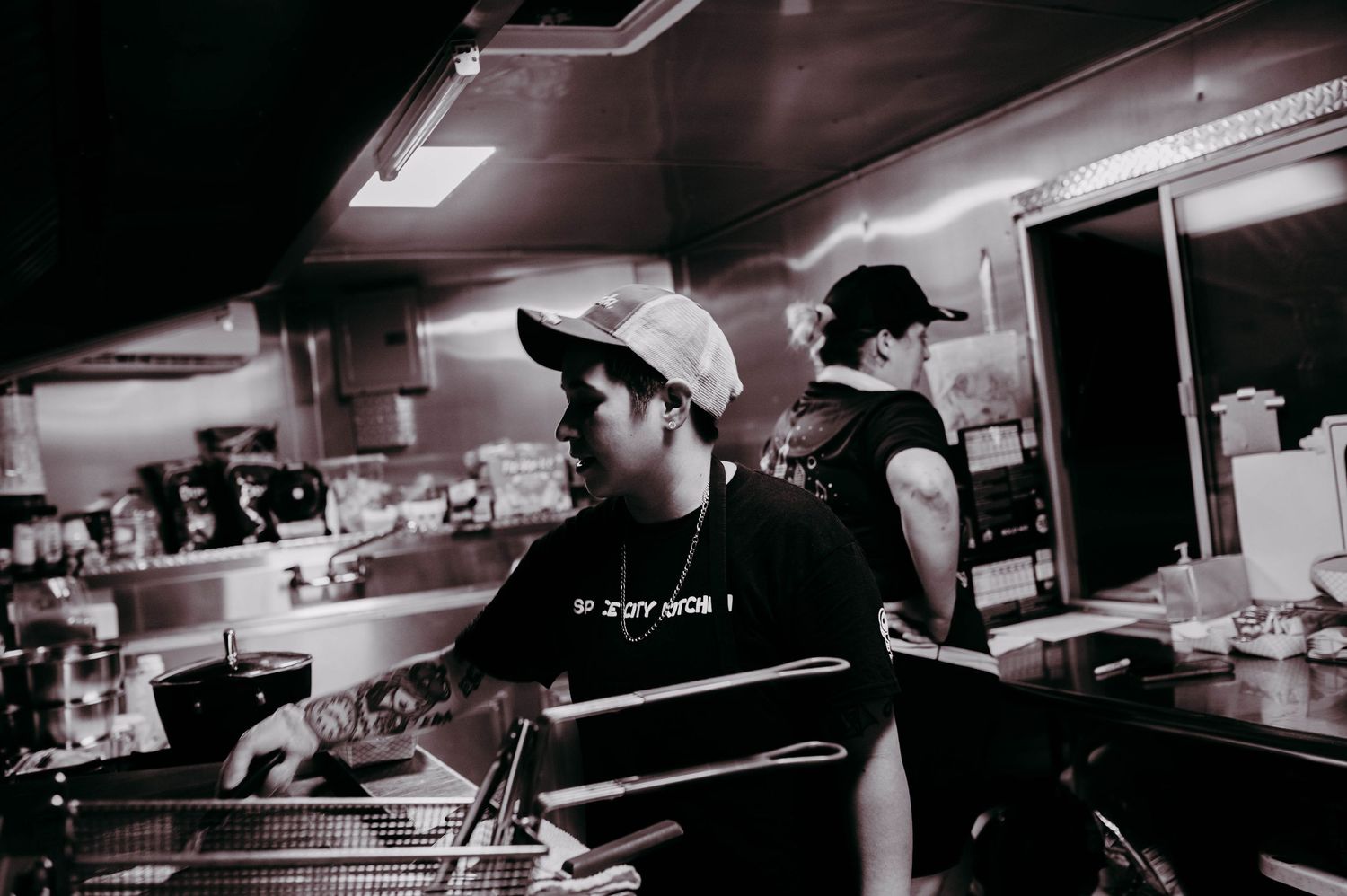 Two cooks inside of a food truck, one who is cooking on a grill and another who is tending to the window.