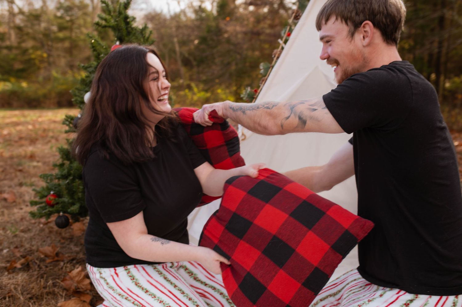 A couple playing with pillows in front of a Christmas tree.