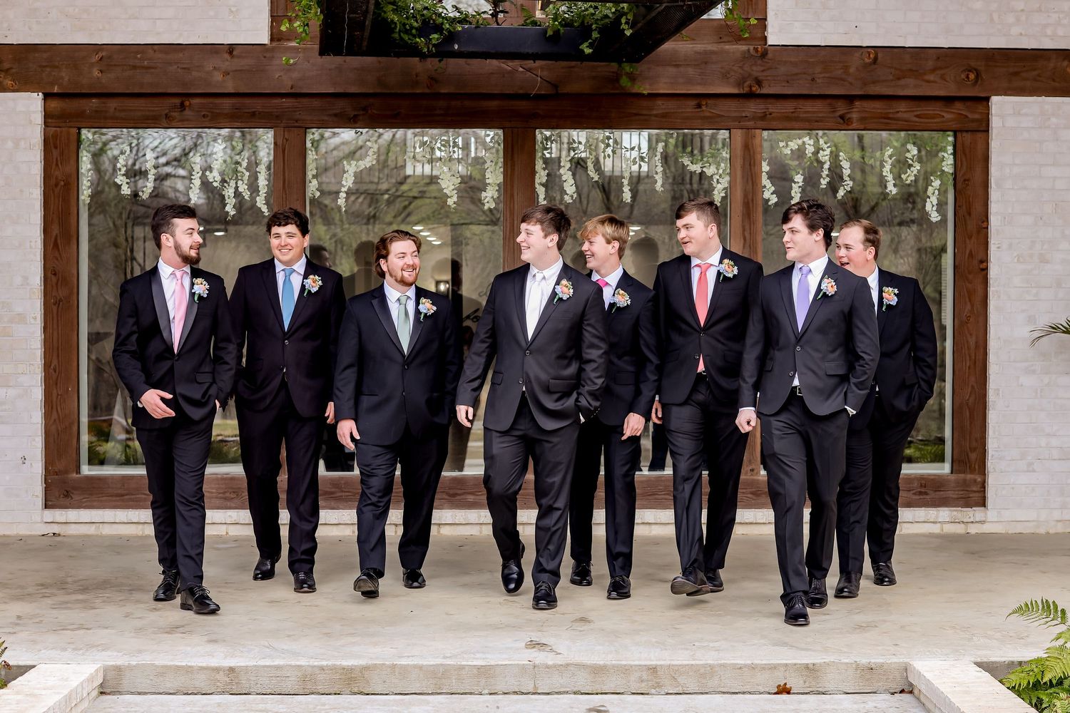 Groomsmen in black suits walking in line along a covered outdoor walkway.