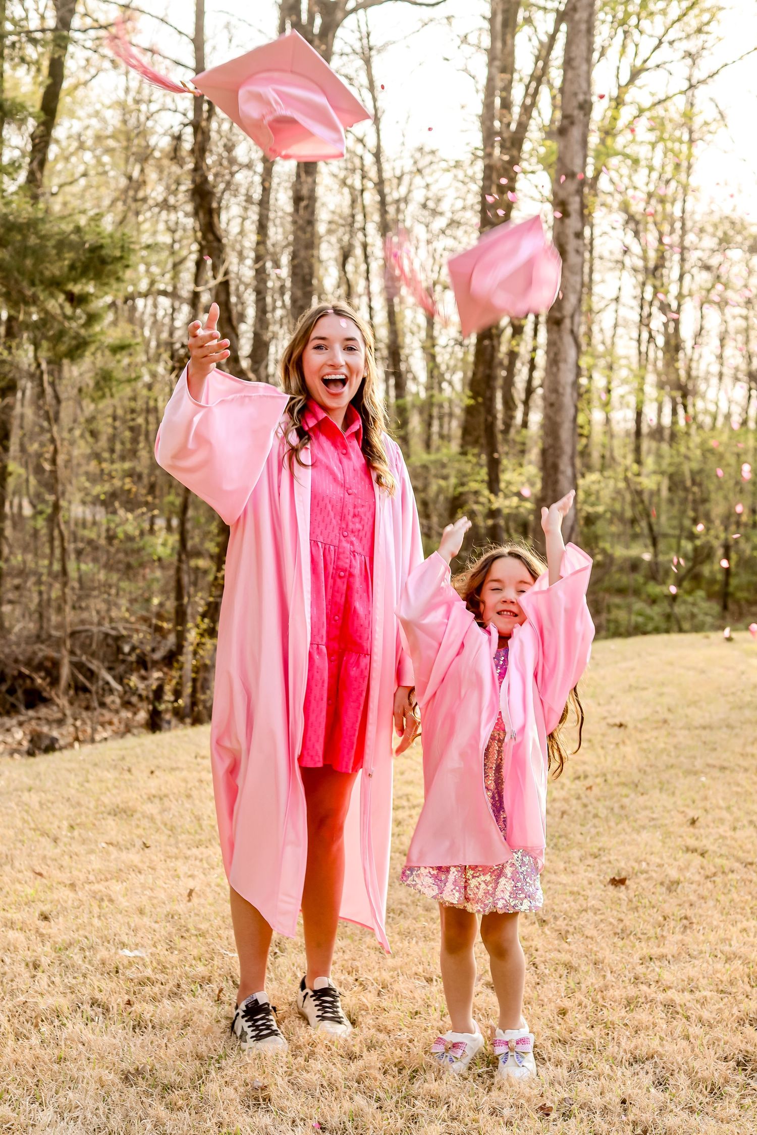 Two graduates in pink robes toss caps joyfully in wooded outdoor setting.