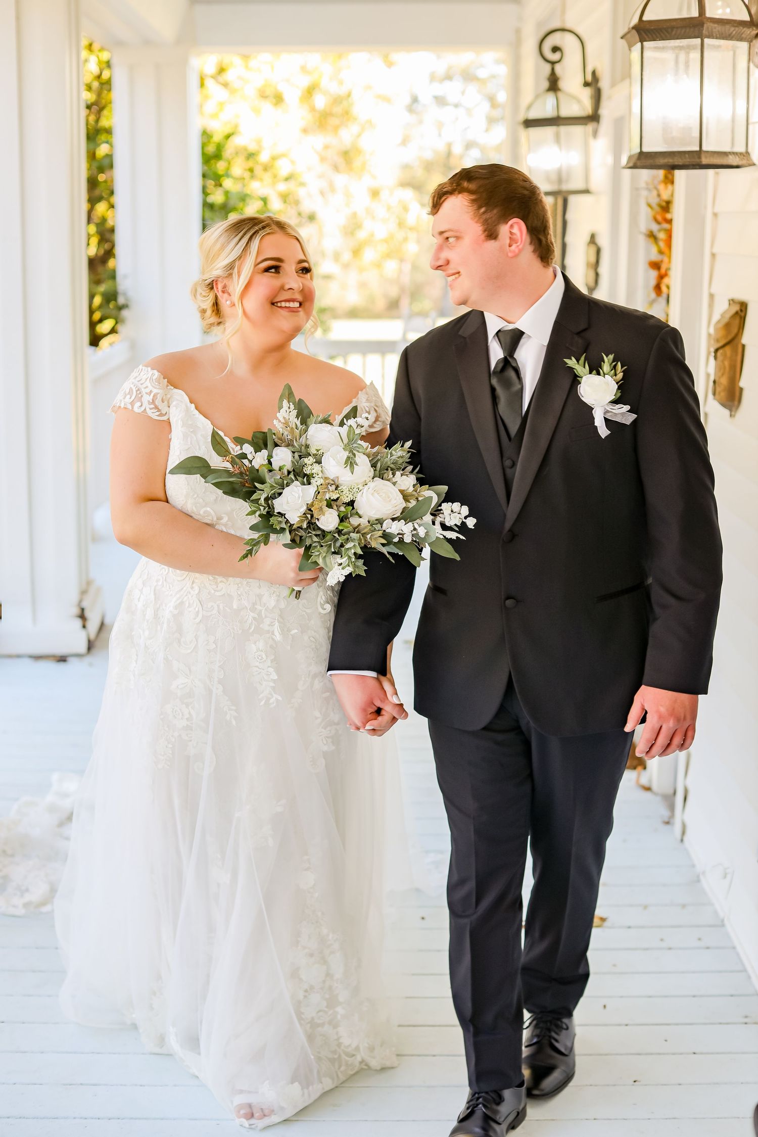 Joyful wedding couple walking and laughing together along a white columned porch with hanging lanterns.