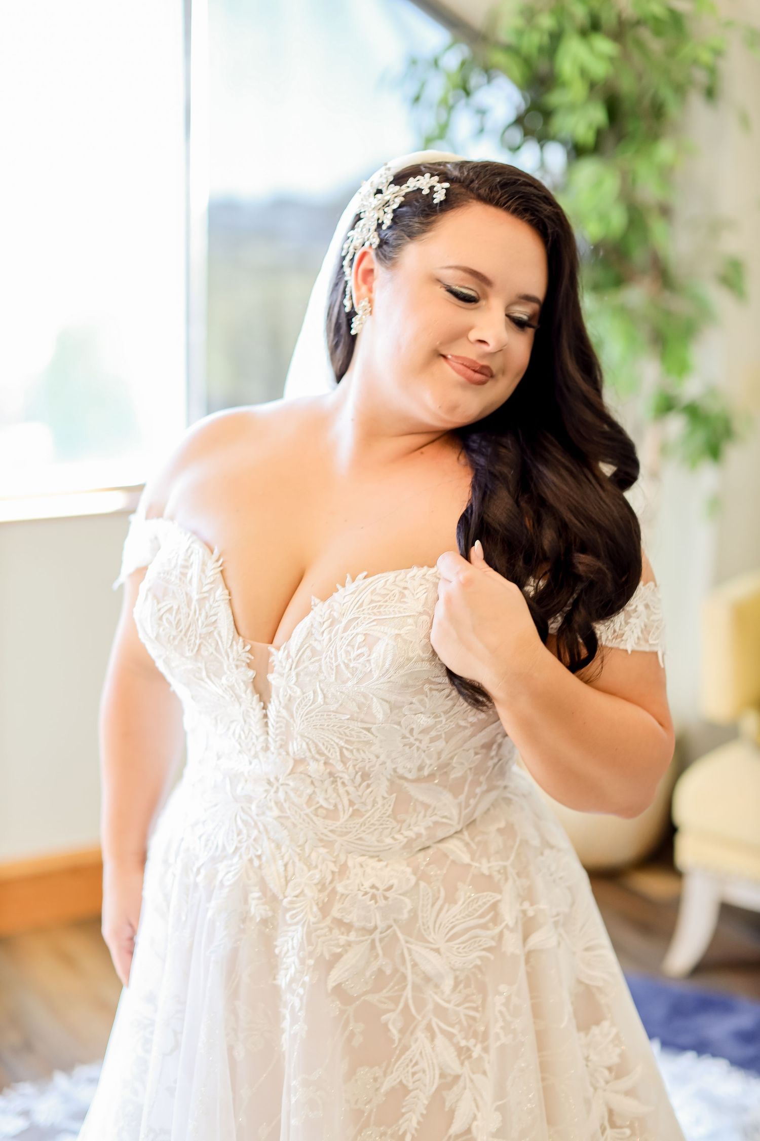 A bride in a detailed lace wedding gown poses elegantly by a window.