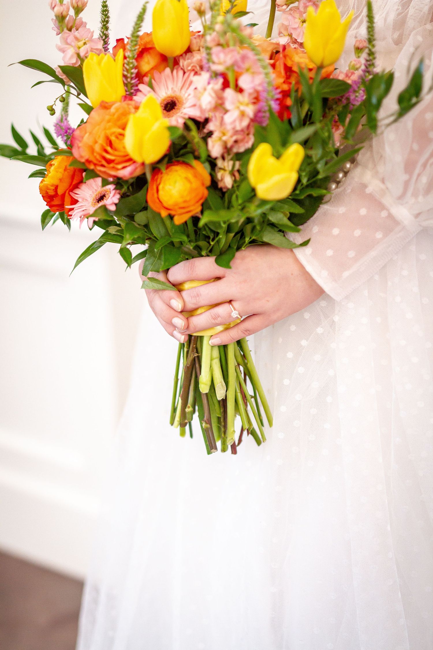 A bright spring wedding bouquet with yellow tulips, orange ranunculus, and pink flowers held against white fabric.