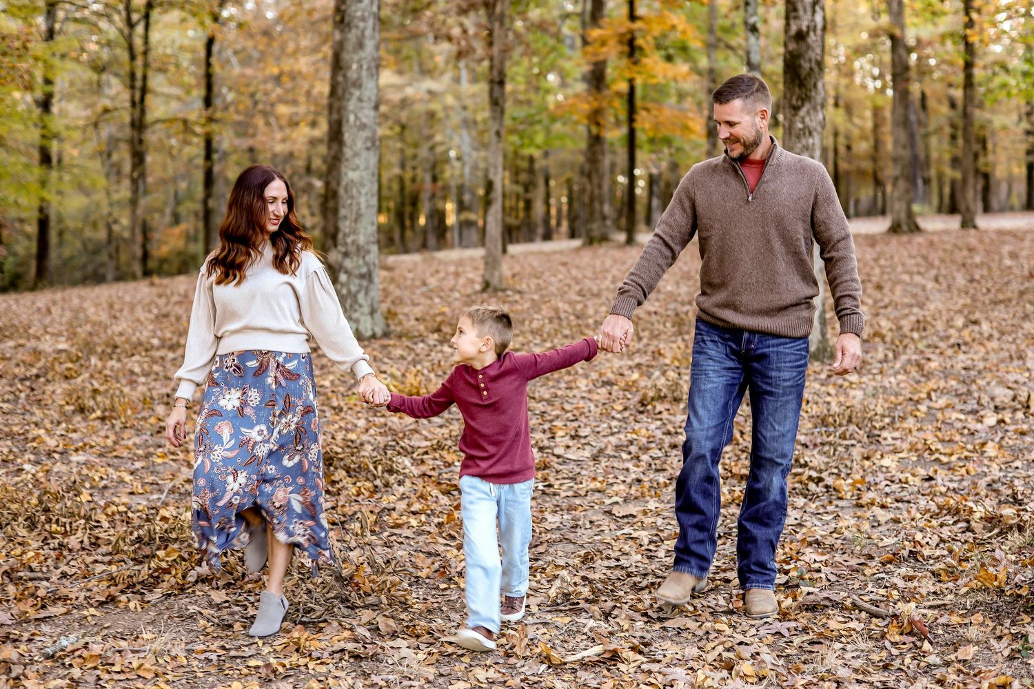 Family walking and playing together on leaf-covered ground in autumn forest setting.