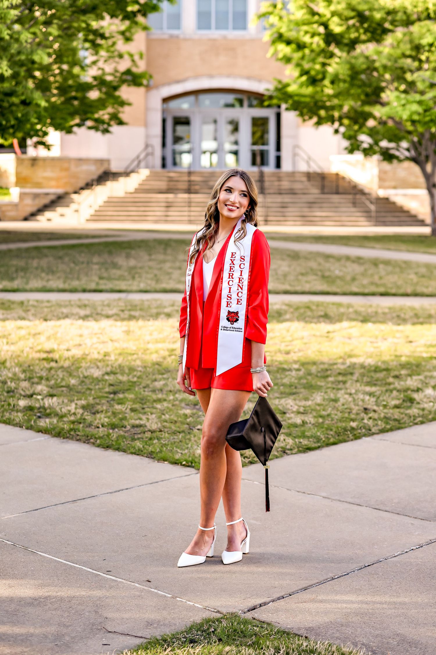 Graduate in red blazer and white sash poses confidently on campus steps with building in background.