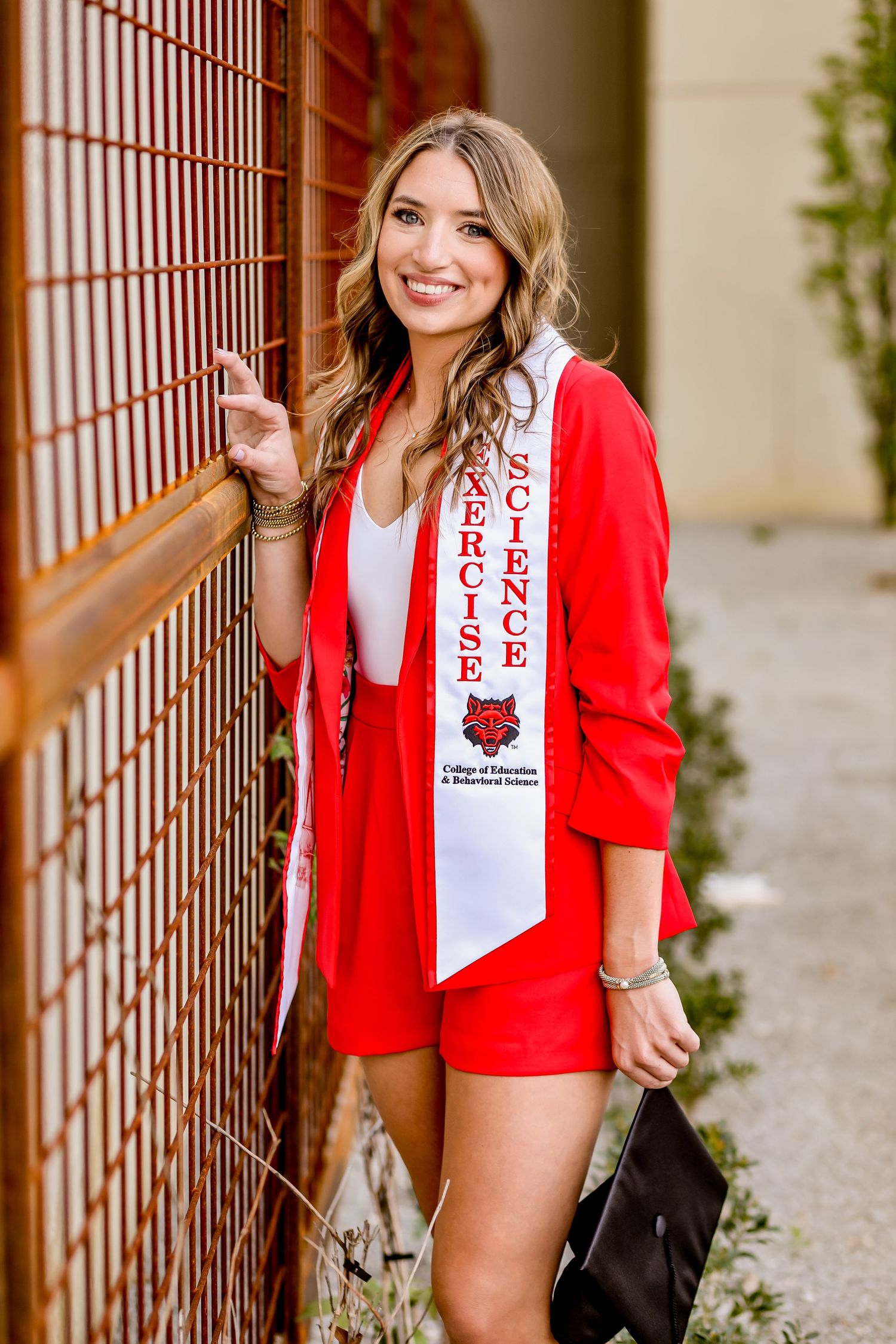 Graduate in red blazer and white sash poses against rustic fence during outdoor photo session.