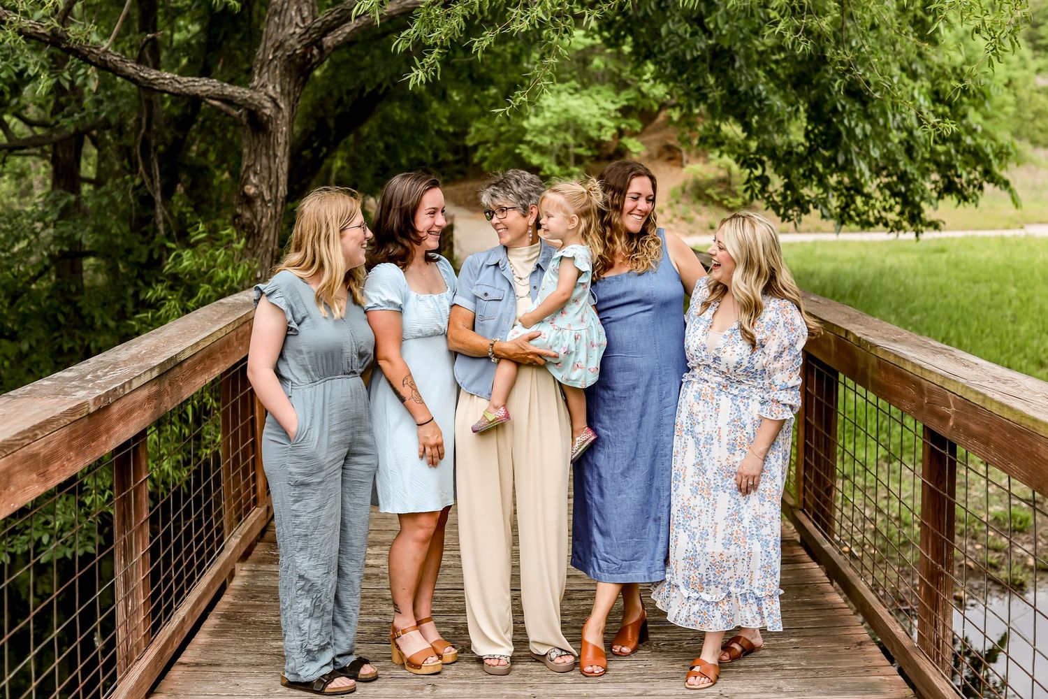 Group of women in coordinating pastel dresses gather on wooden bridge for photo.