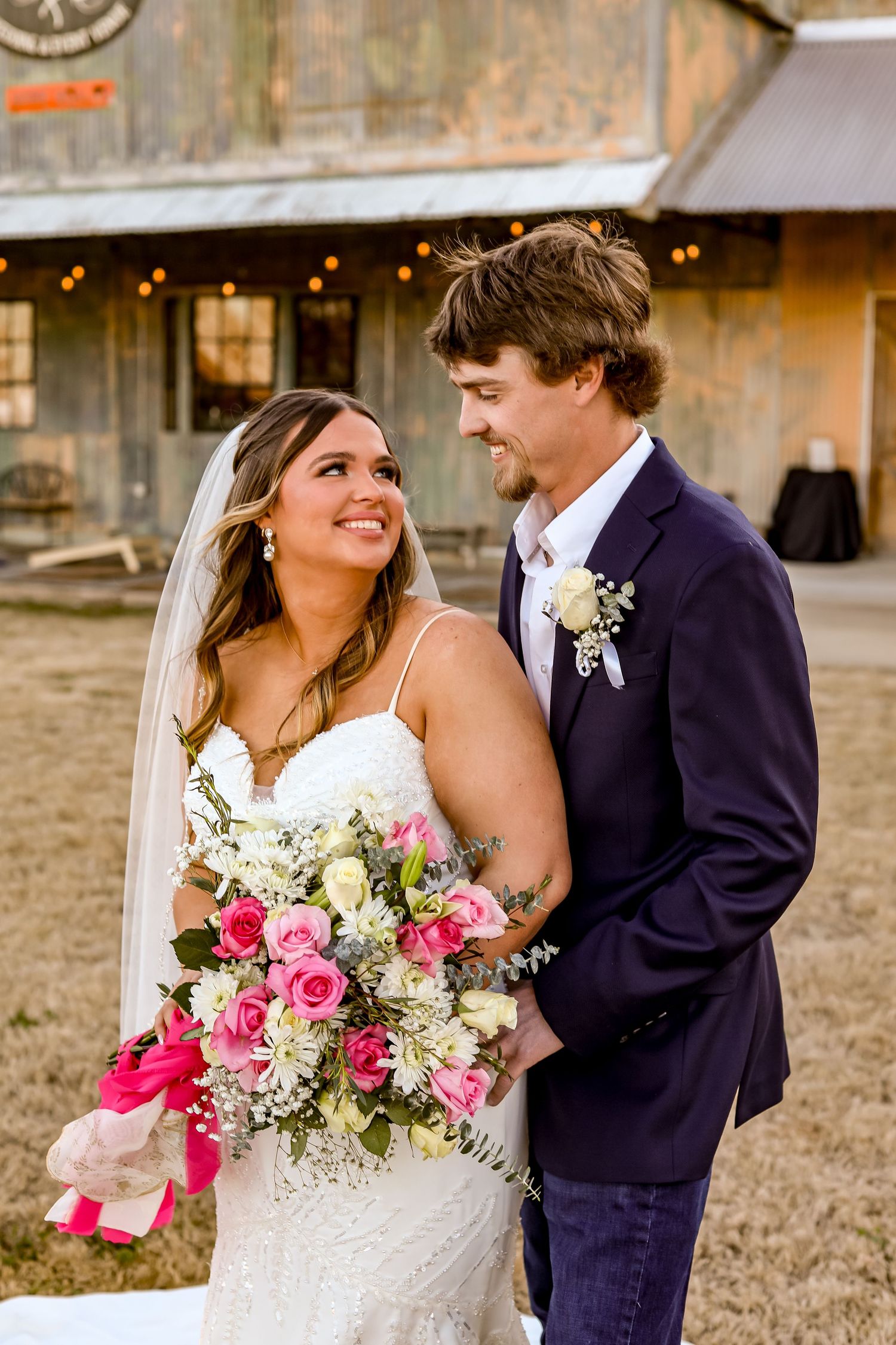 A joyful wedding couple shares a tender moment outside a rustic barn venue at sunset.
