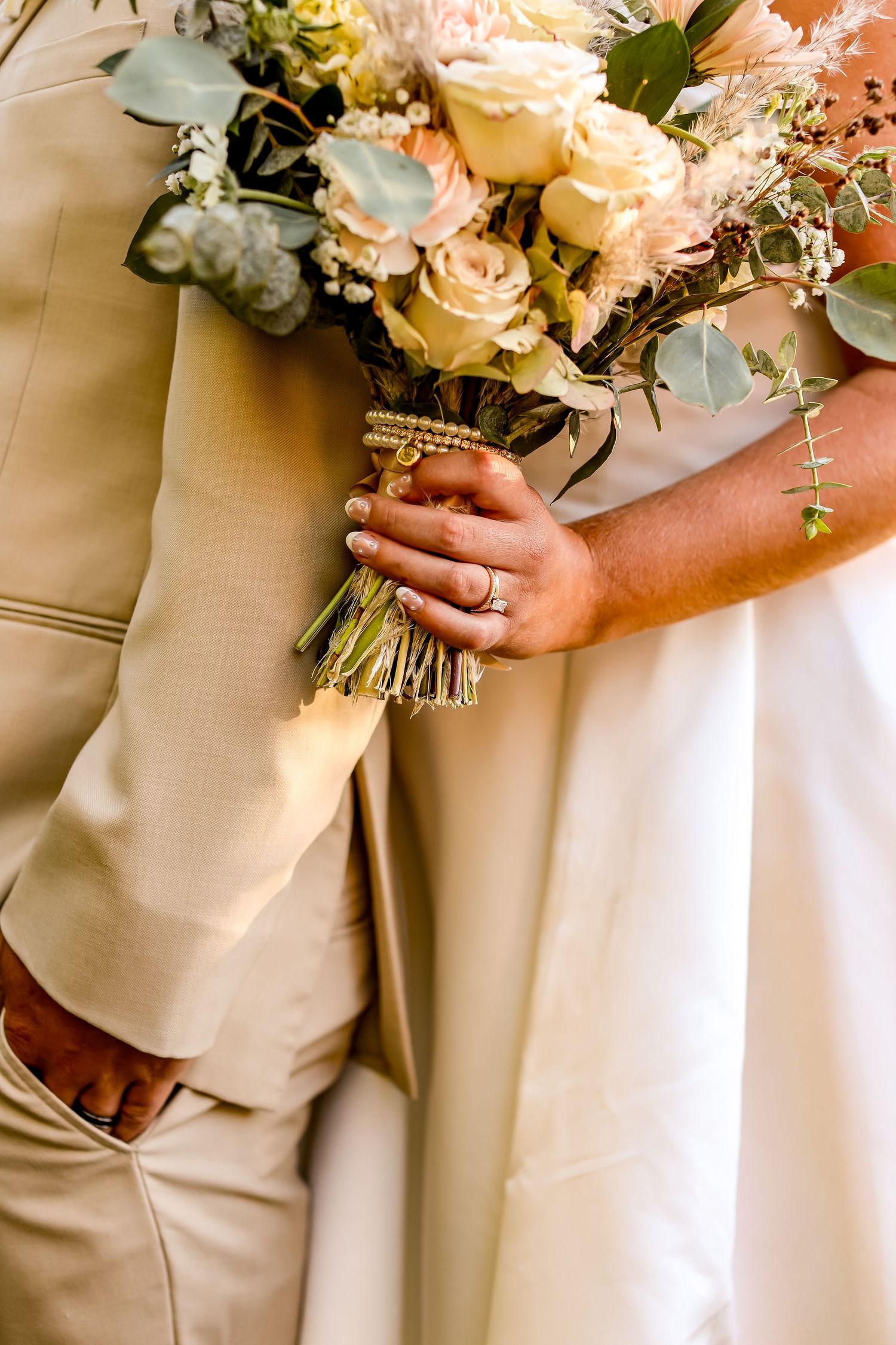Close-up of wedding bouquet with blush roses and eucalyptus held against beige and white formal attire.