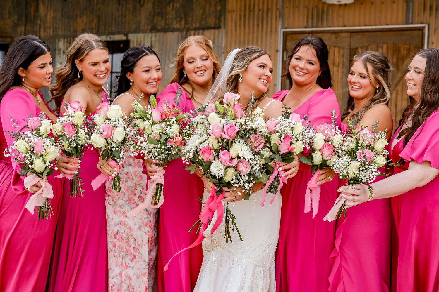 A bridal party in bright pink dresses holds matching floral bouquets while laughing together.