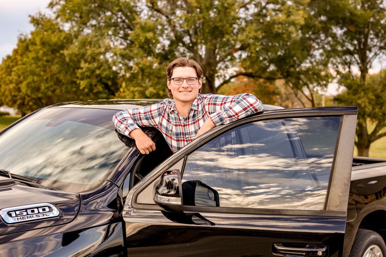 Person in plaid shirt poses happily while leaning on black car outdoors.