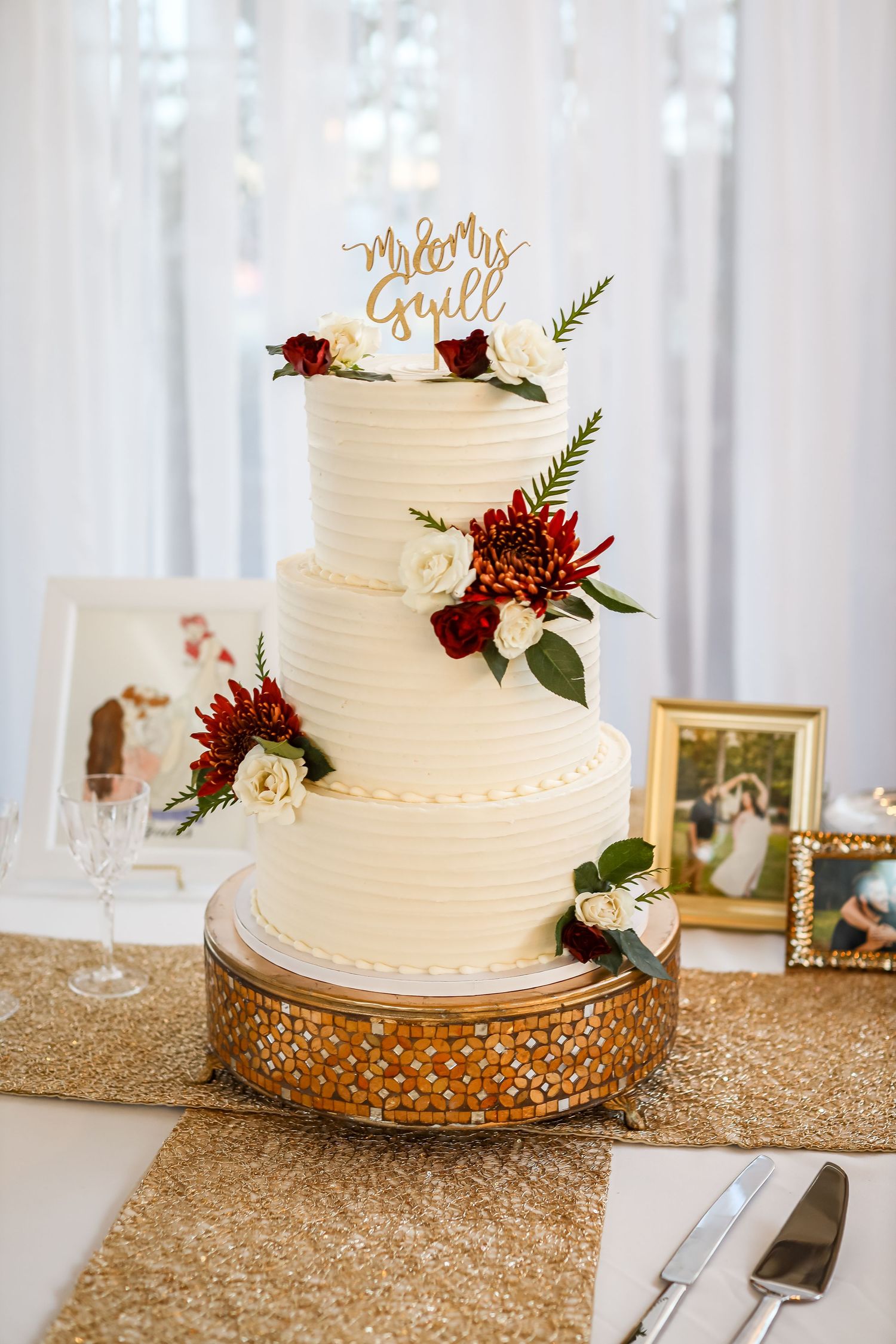 An elegant three-tier white wedding cake decorated with burgundy flowers on a gold sequin tablecloth.