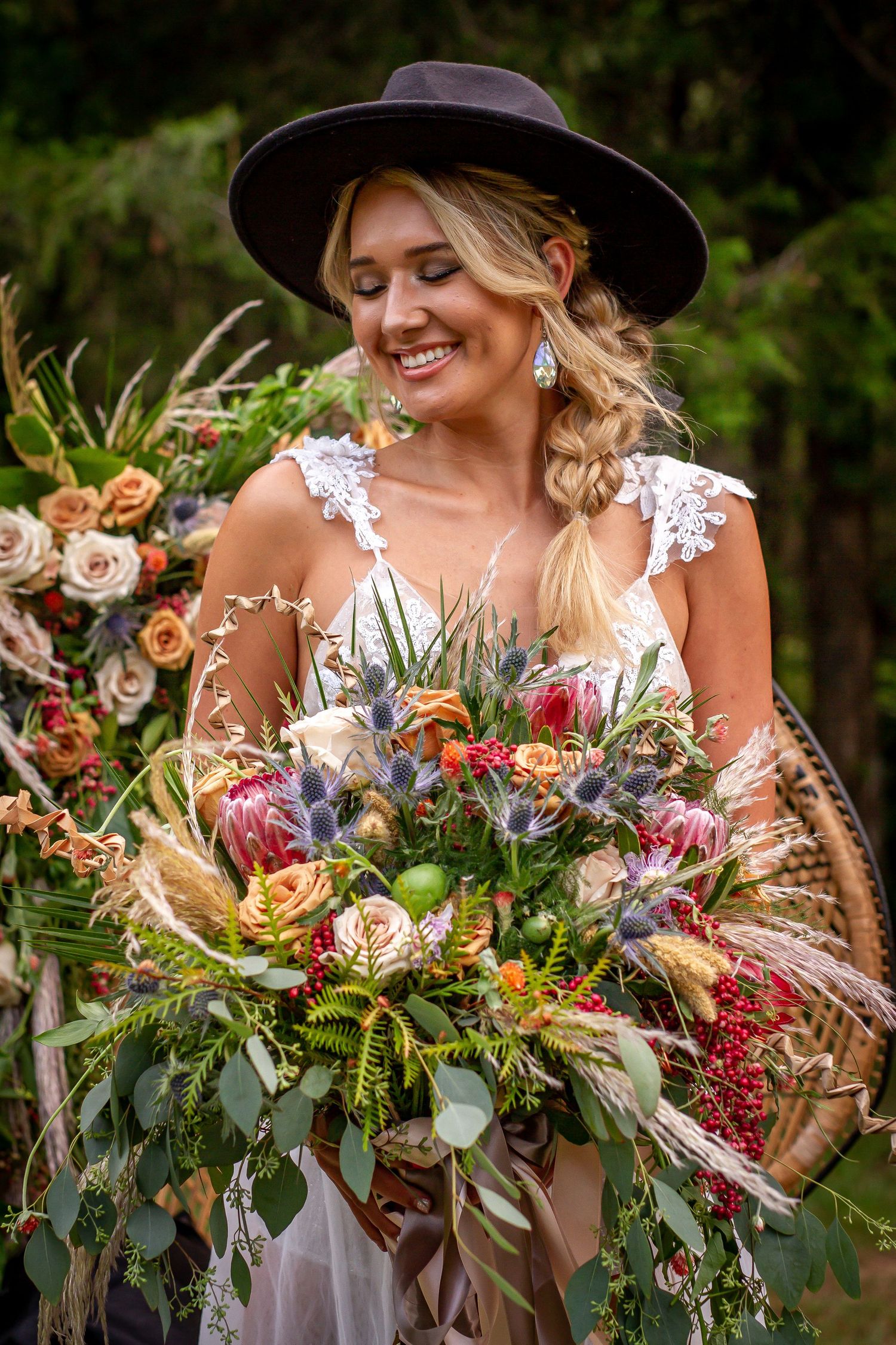 A boho bride wearing a wide-brimmed hat holds a wild, colorful bouquet with dried elements and roses.