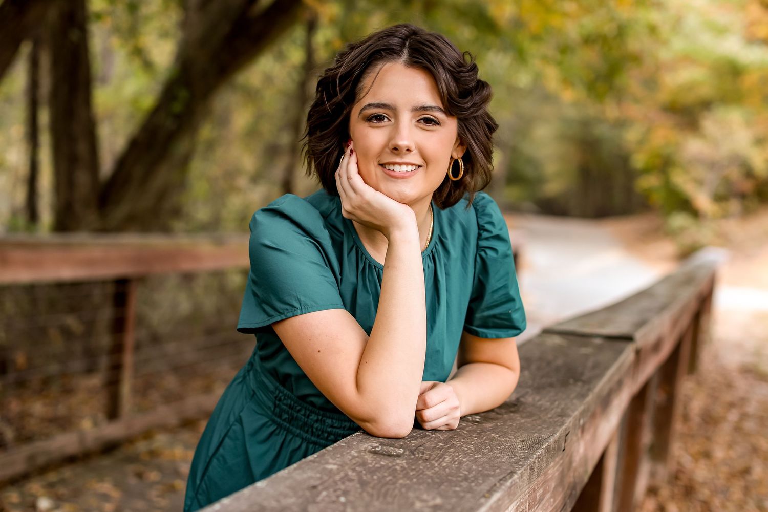 Portrait of someone in teal shirt leaning on wooden railing in autumn setting.