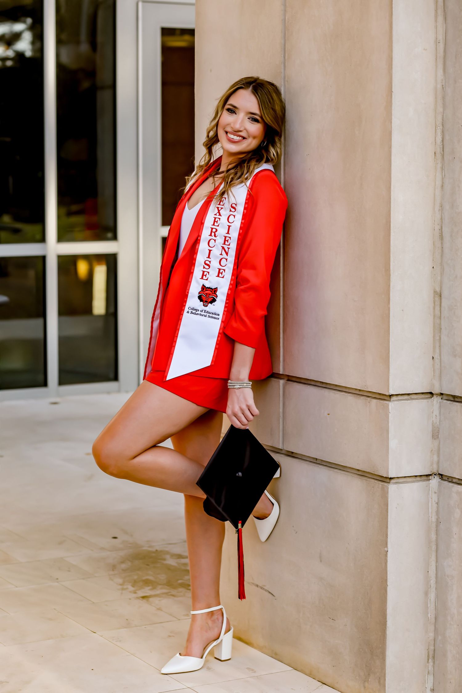 Graduate in red stole poses against stone building during graduation photoshoot.