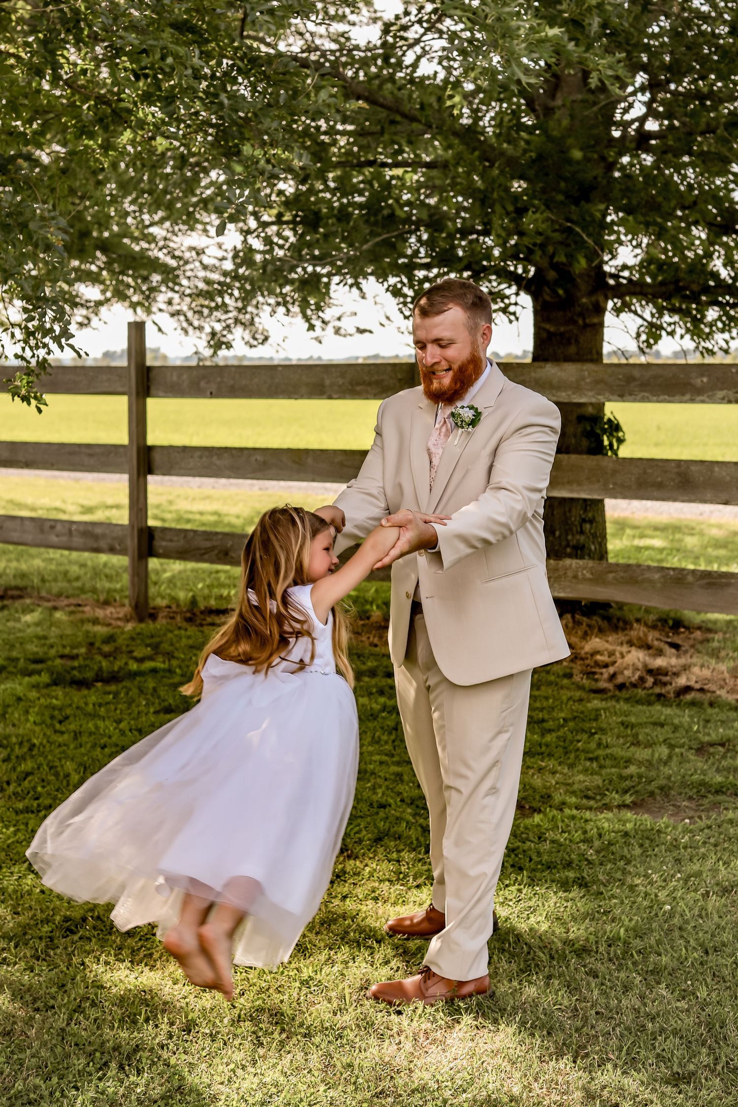 Father in light beige suit dances with daughter in white dress on grassy lawn.