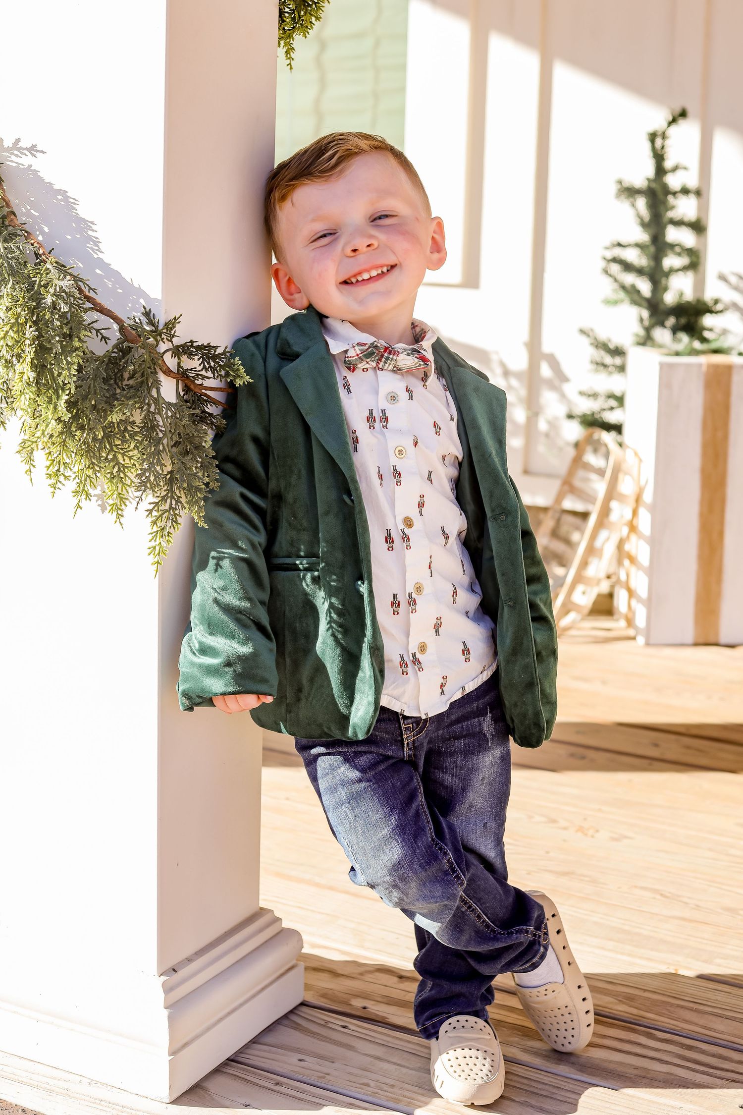 Stylishly dressed young child in green blazer and jeans poses on sunny porch with decorative greenery.