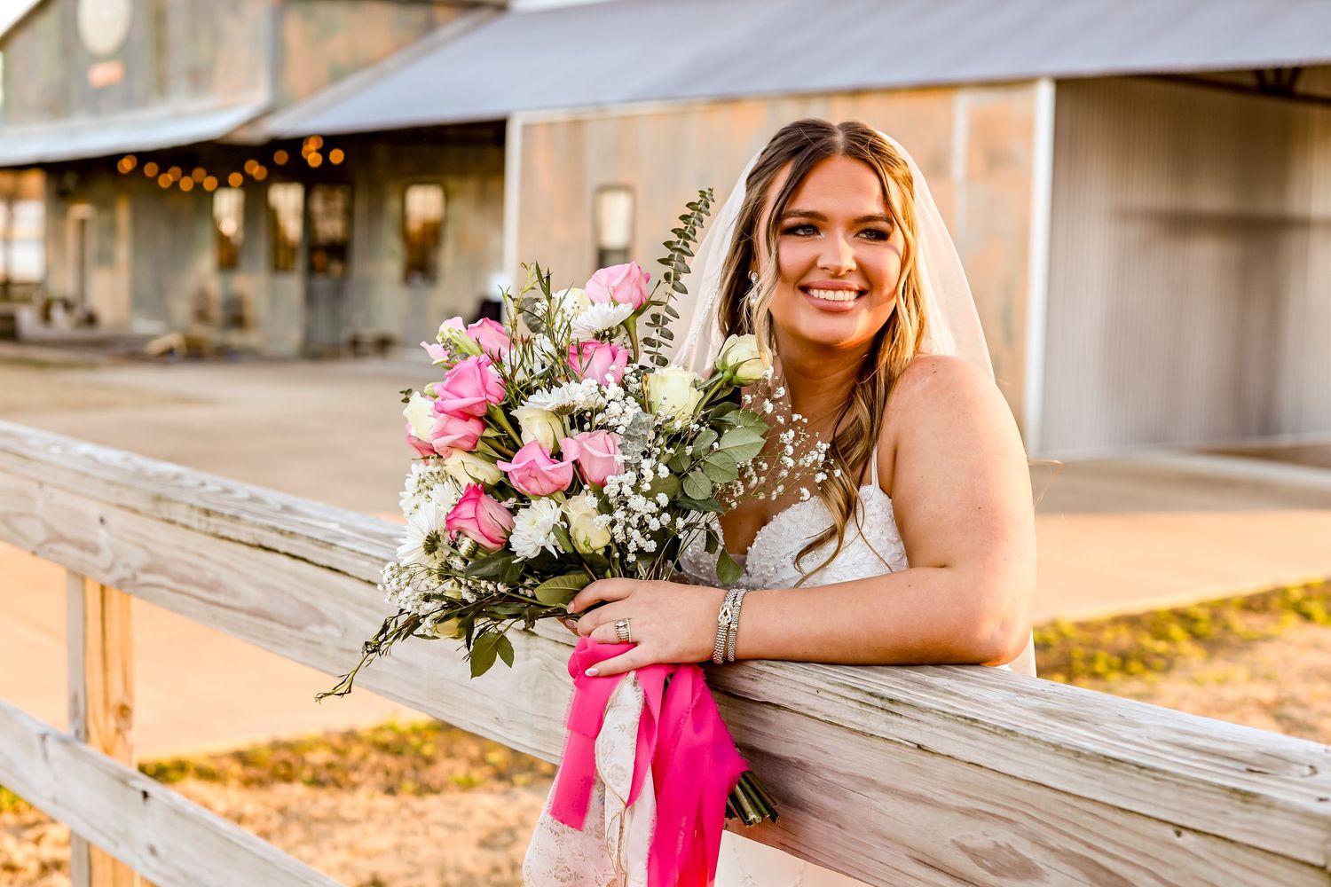 A bride in a white dress poses with a vibrant pink and white bouquet against a rustic fence.
