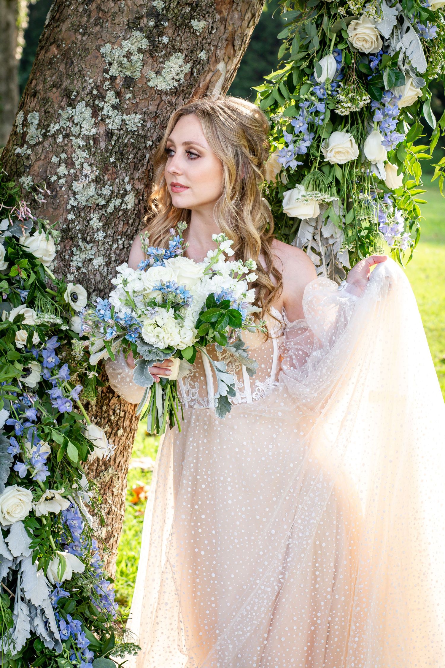A bride in a lace dress holds a blue and white bouquet against a tree decorated with floral arrangements.