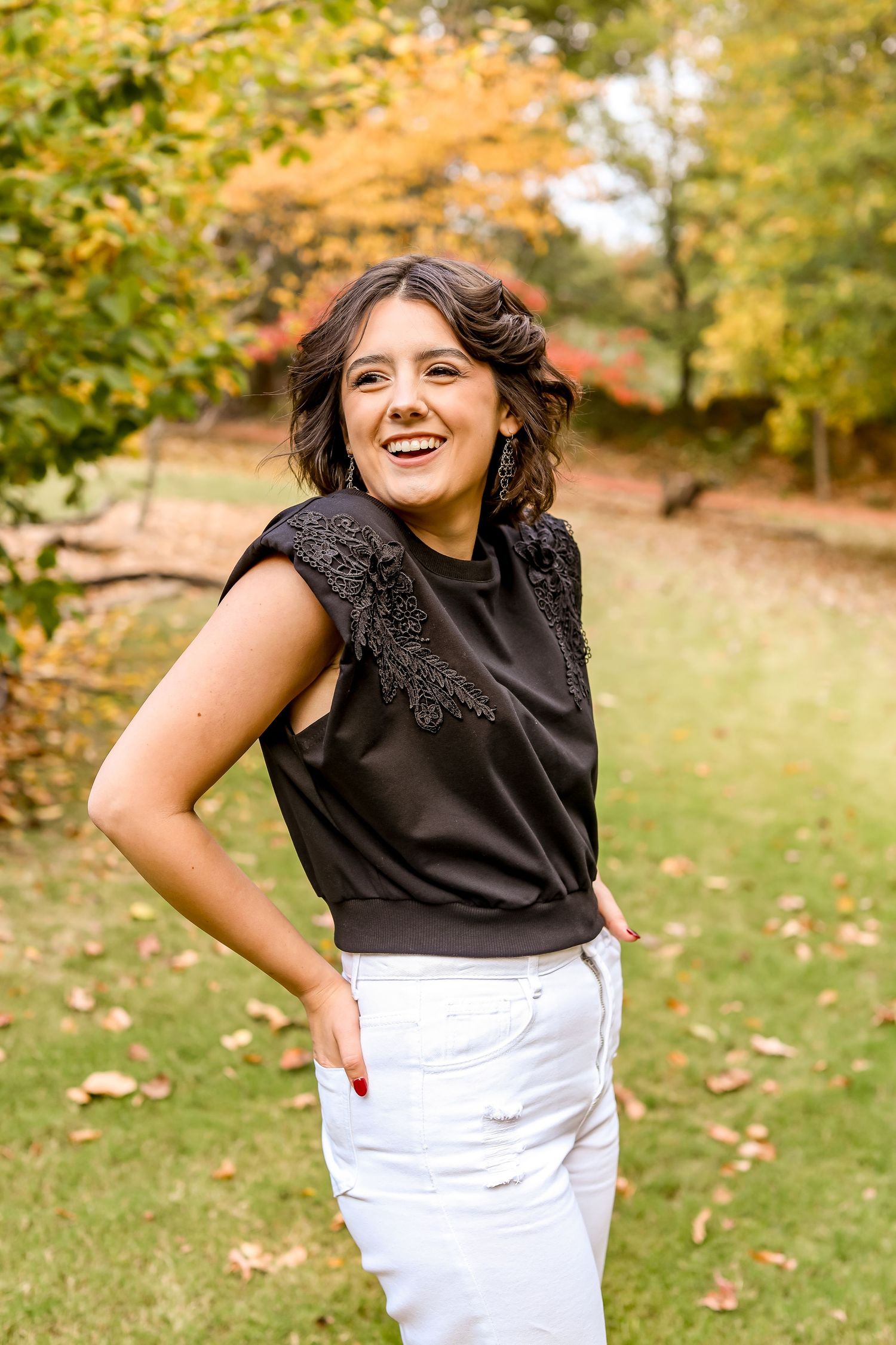 Portrait in black lace top and white pants against autumn trees.