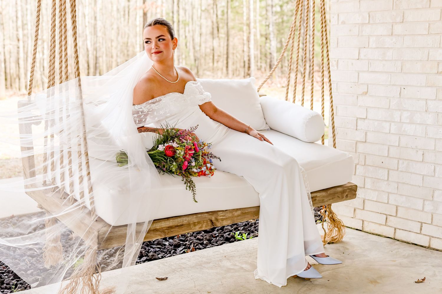 Bride in flowing white dress poses against elegant white brick wall with string lights.