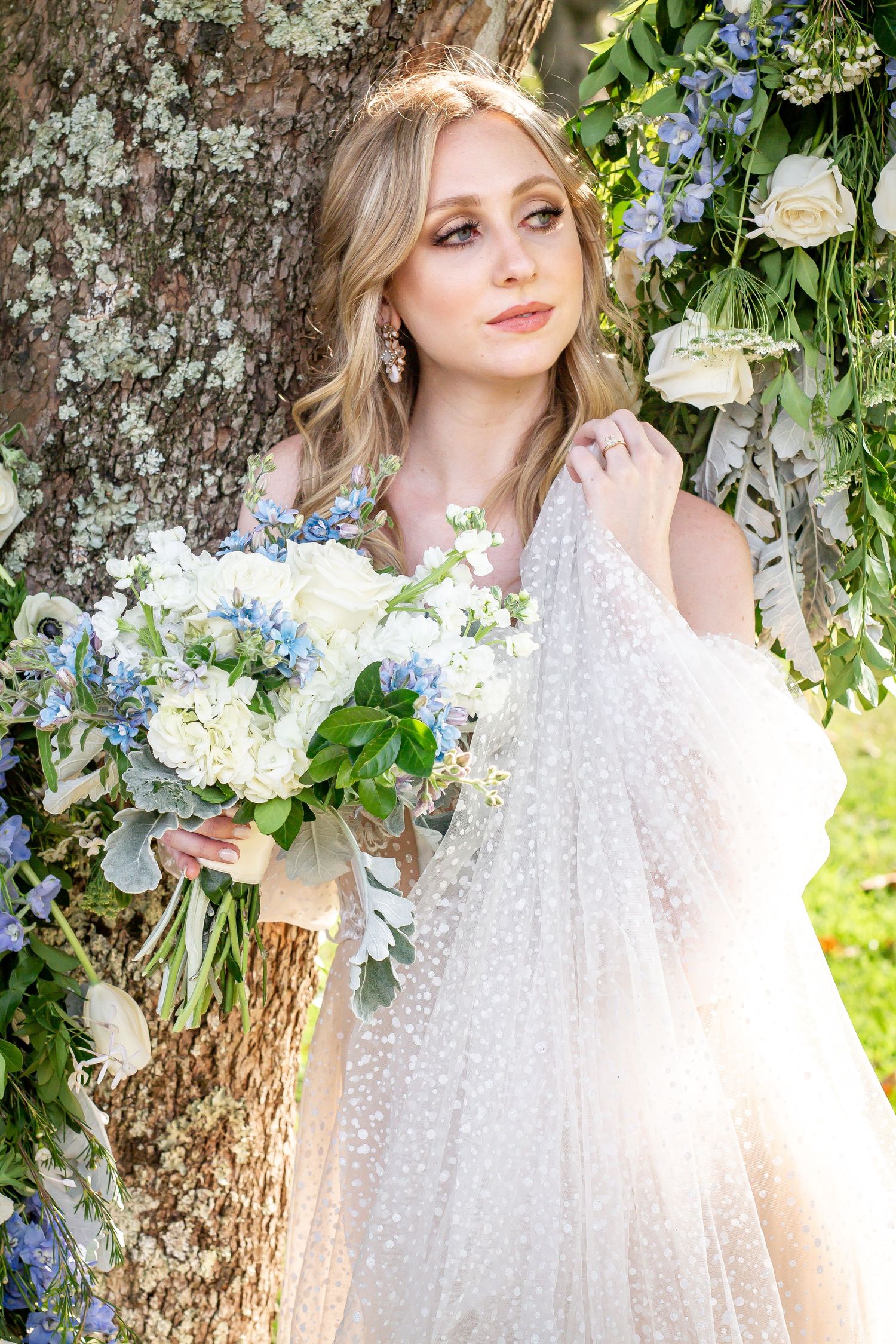Ethereal bridal portrait with blue and white flowers against a mossy tree trunk.