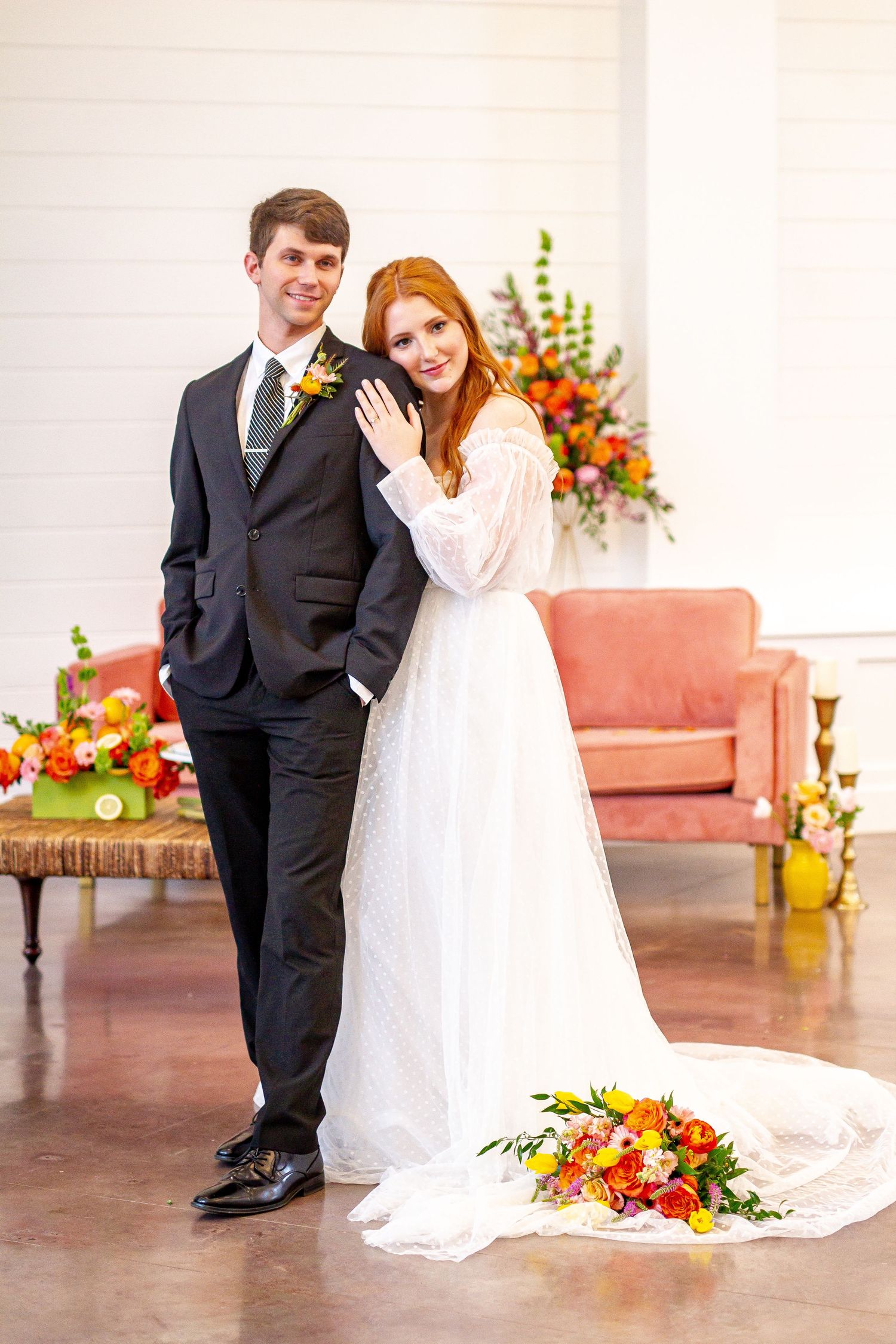 A wedding couple poses in front of vibrant orange and yellow floral arrangements and a pink velvet couch.