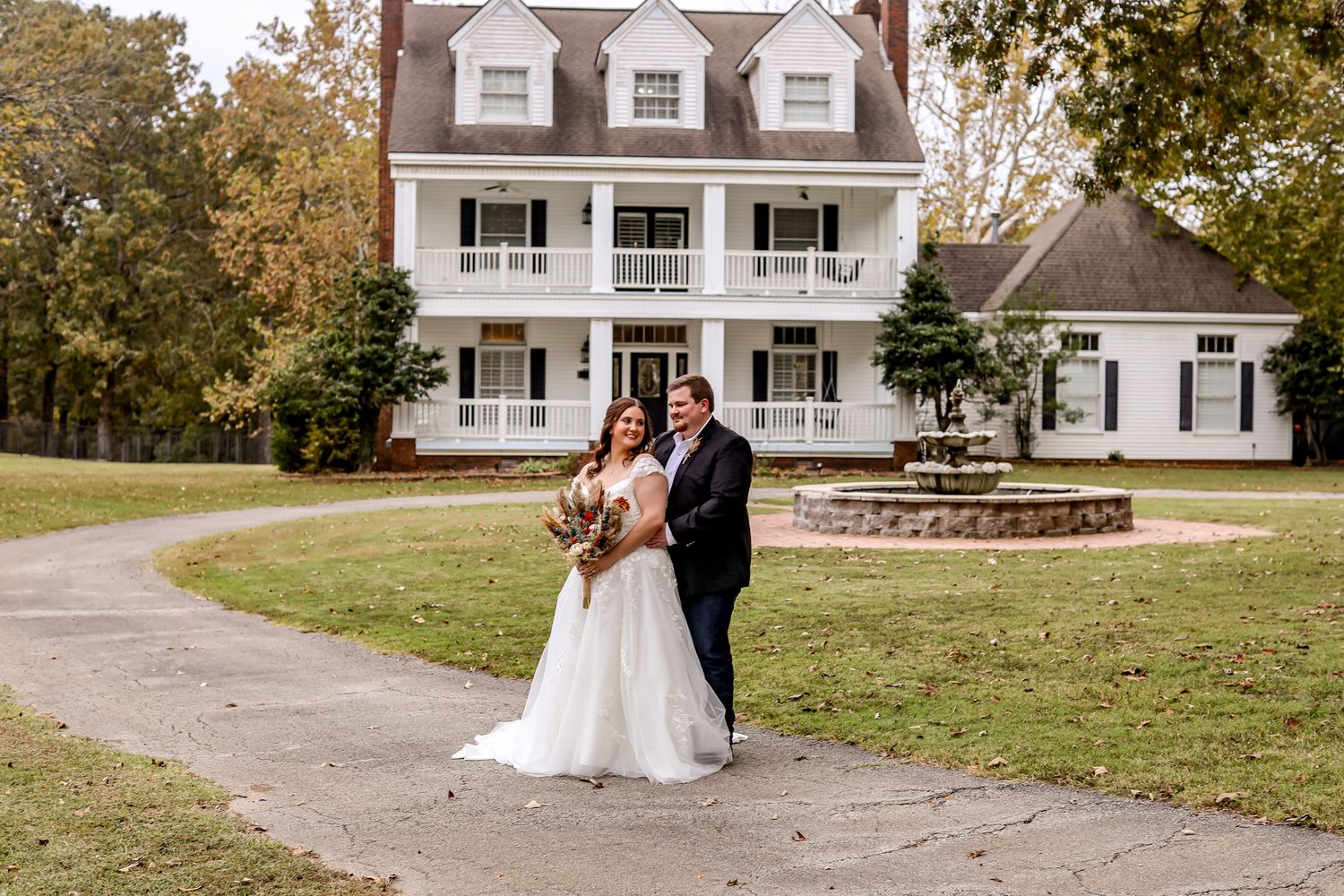 Newlyweds pose in front of a classic white colonial style house with dormer windows.