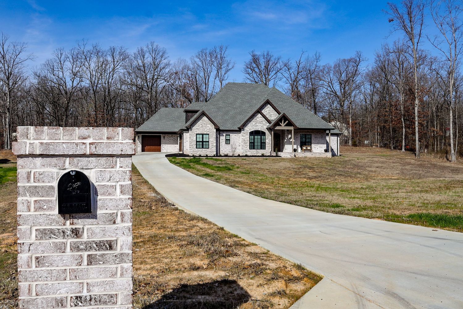 Long driveway leading to modern farmhouse with stone accents and surrounding trees.