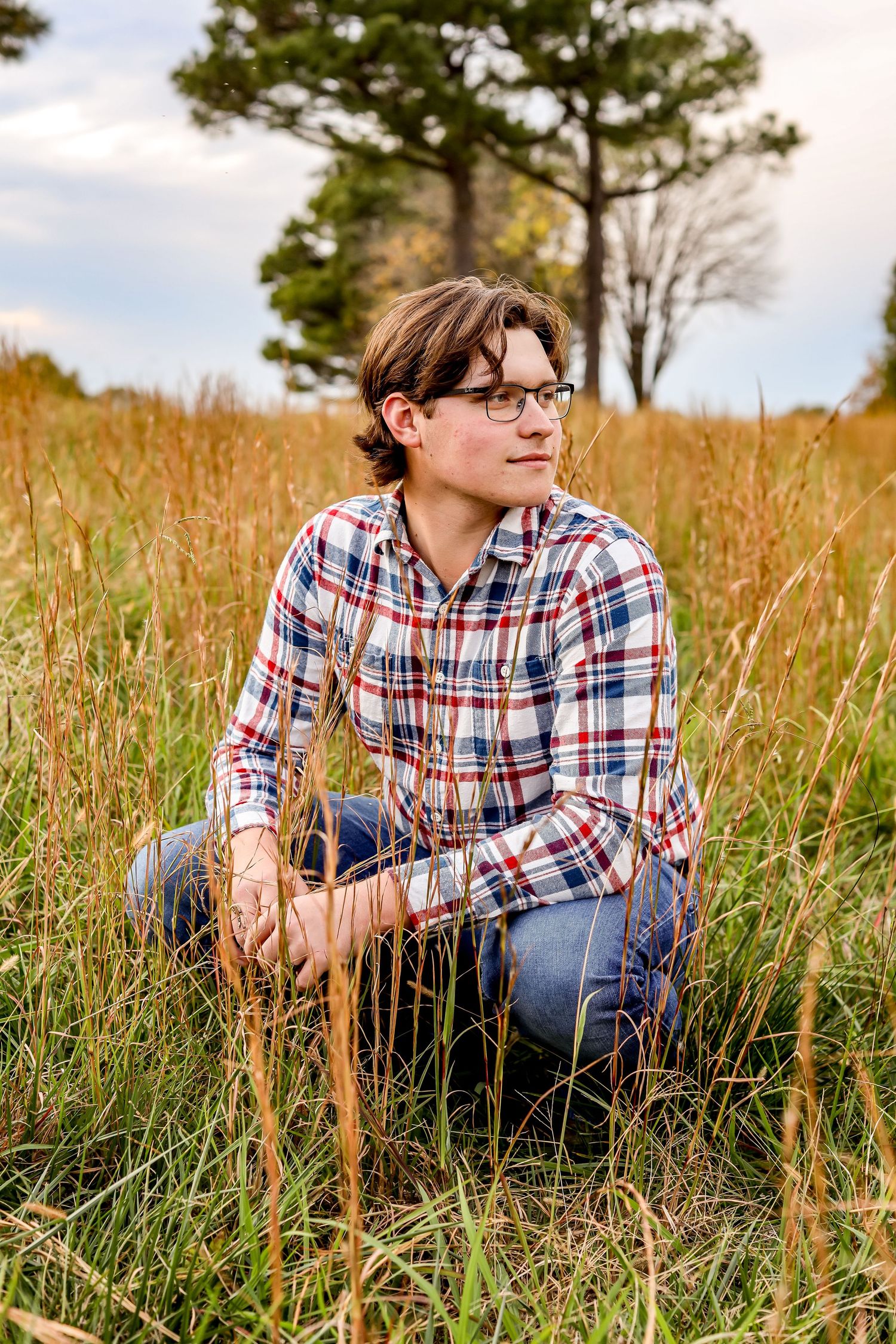 Person in plaid shirt and glasses kneeling in tall autumn grass with trees in background.