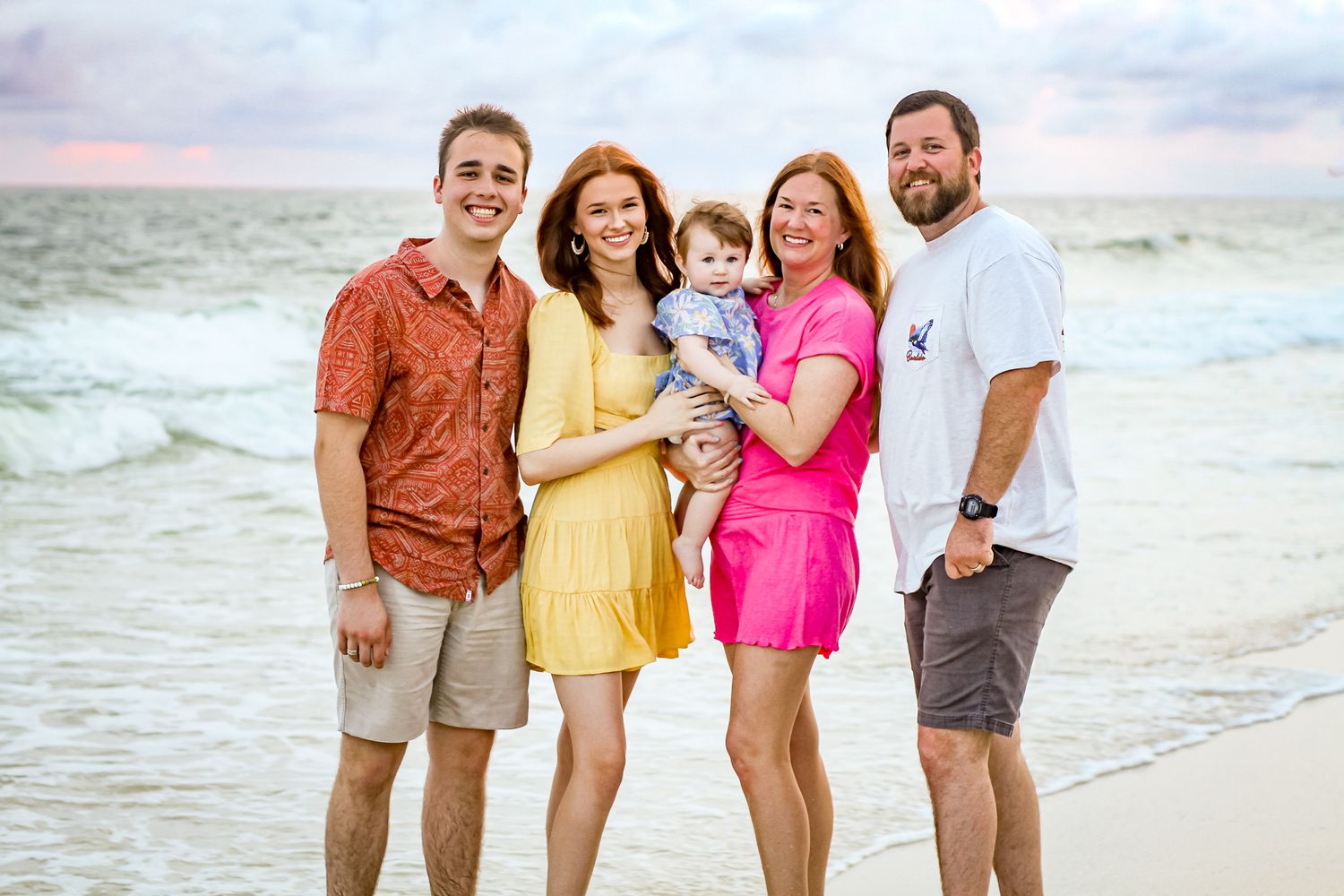 A happy family poses together on a sunny beach wearing coordinated yellow and pink outfits.