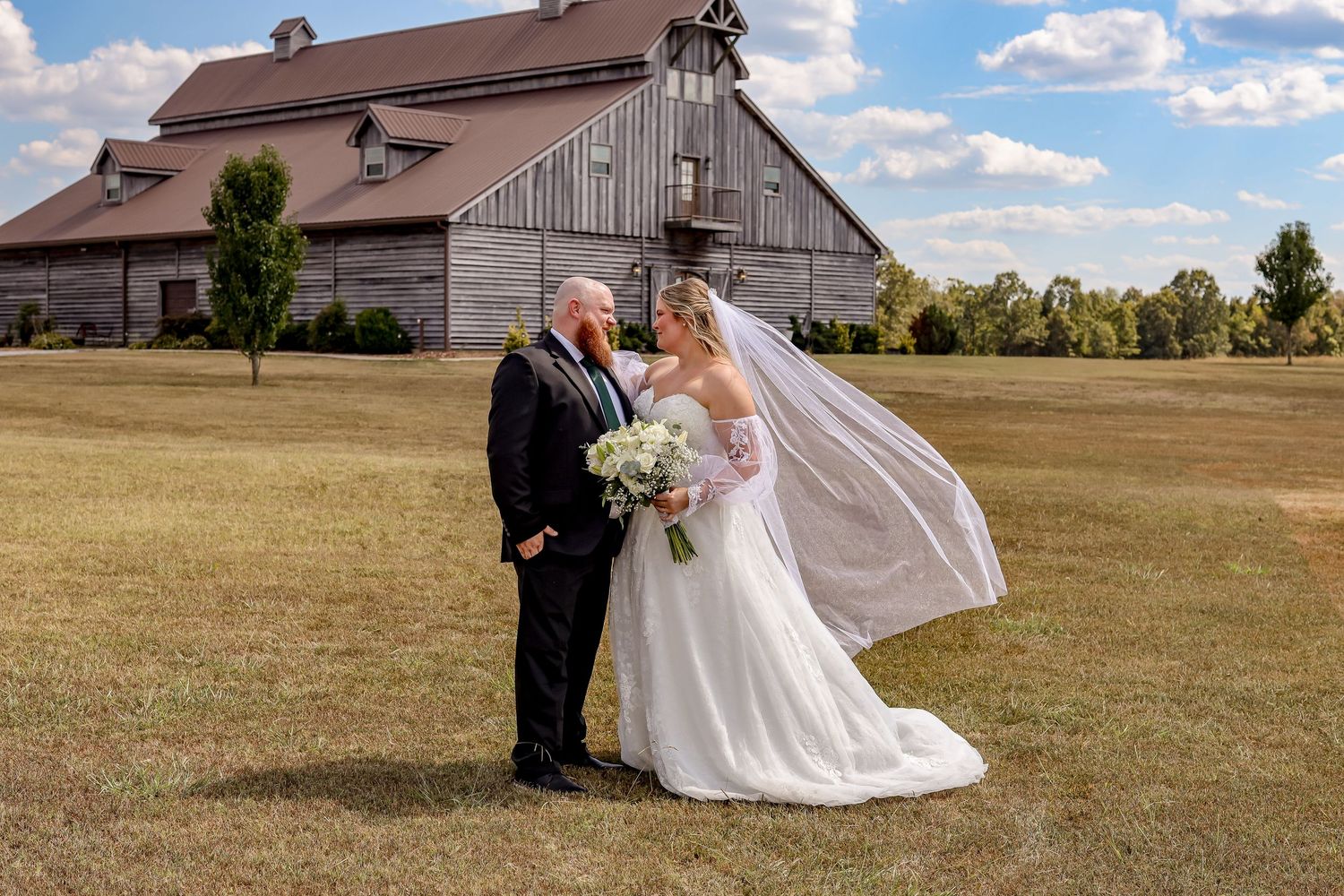 Rustic barn wedding photos with couple posing in front of weathered wooden buildings in an open field.