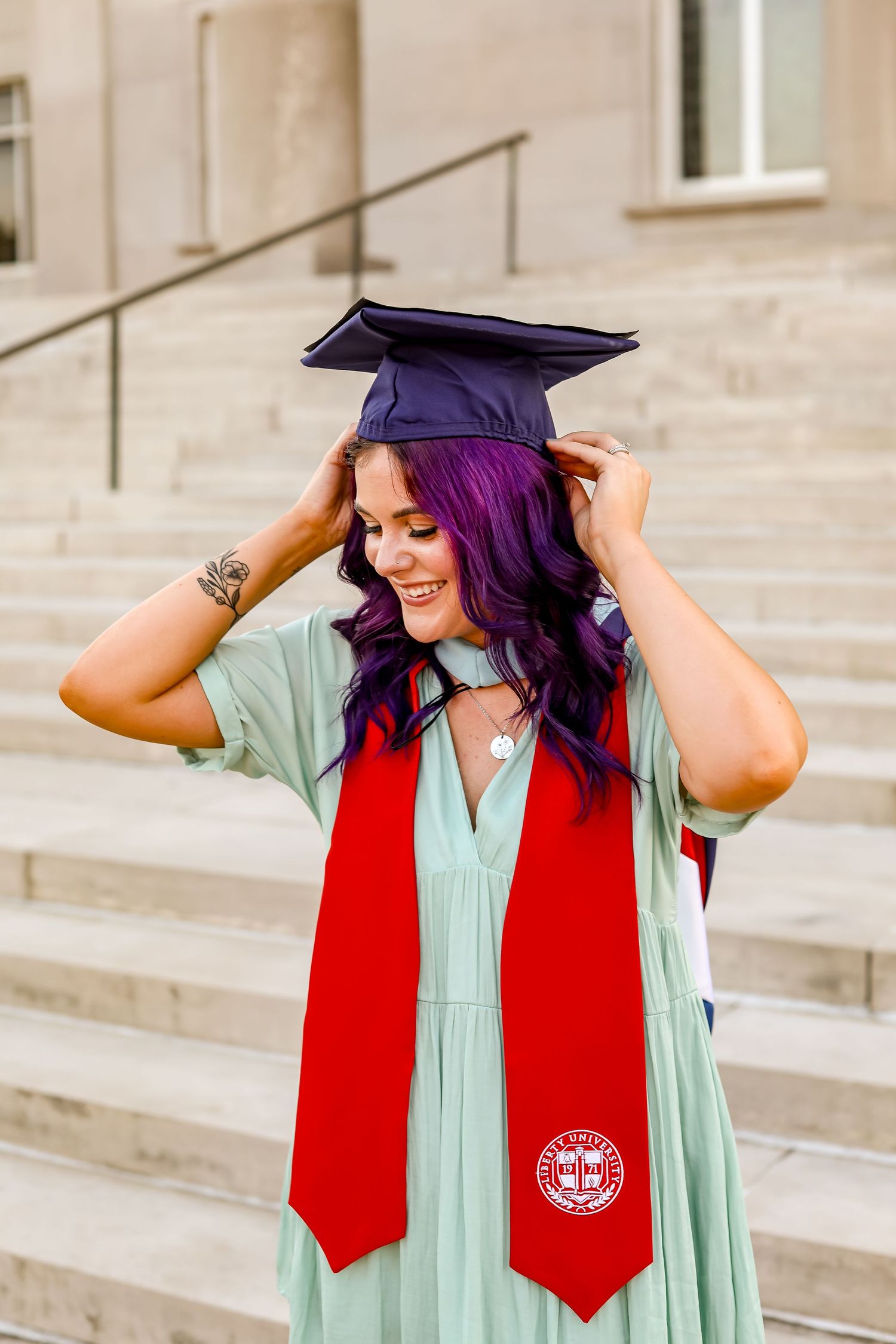Graduate adjusts cap while wearing mint green gown and red stole on stone steps.