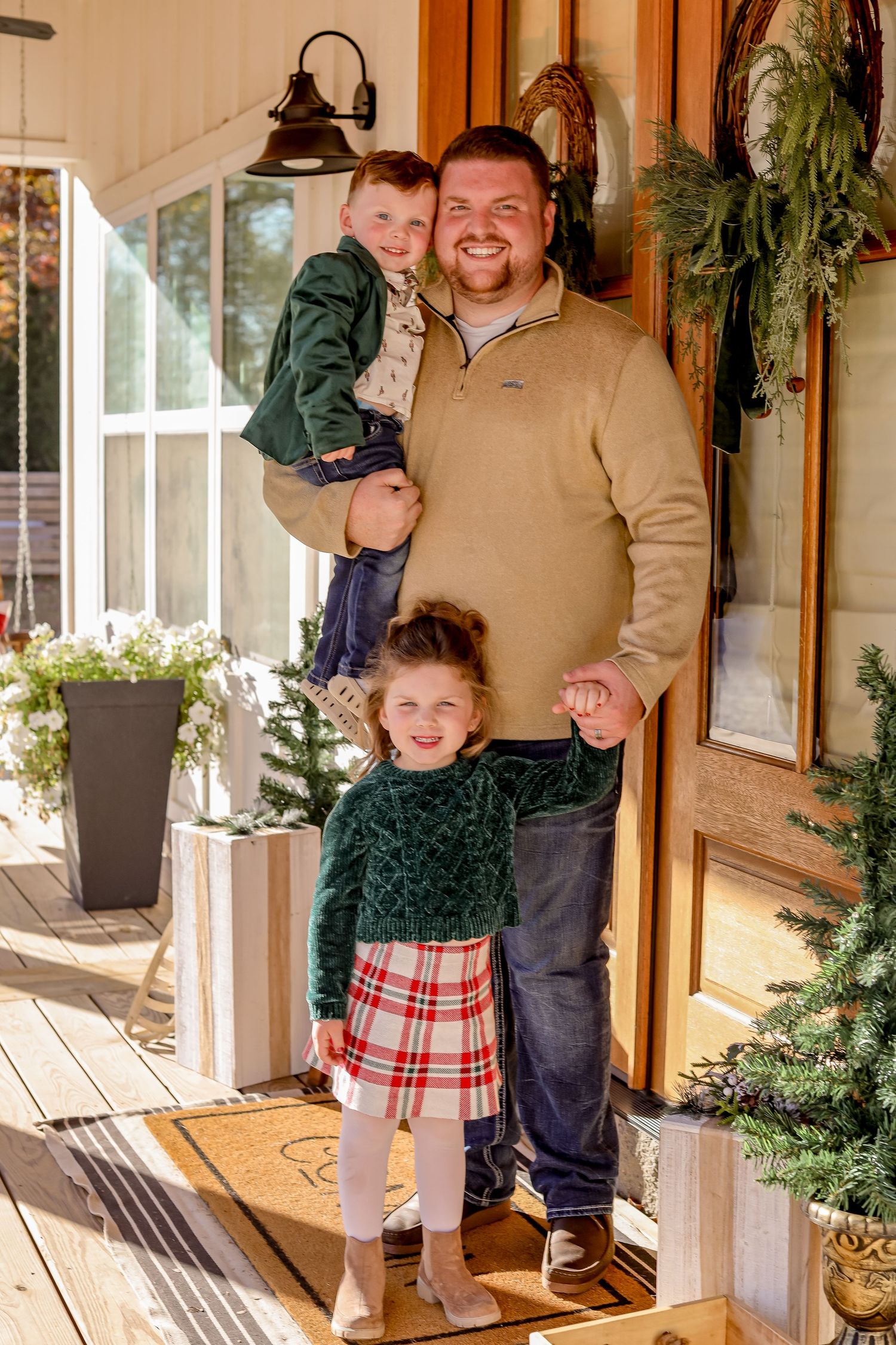 A smiling father holds his young son while his daughter stands in front on a festive porch decorated for Christmas.