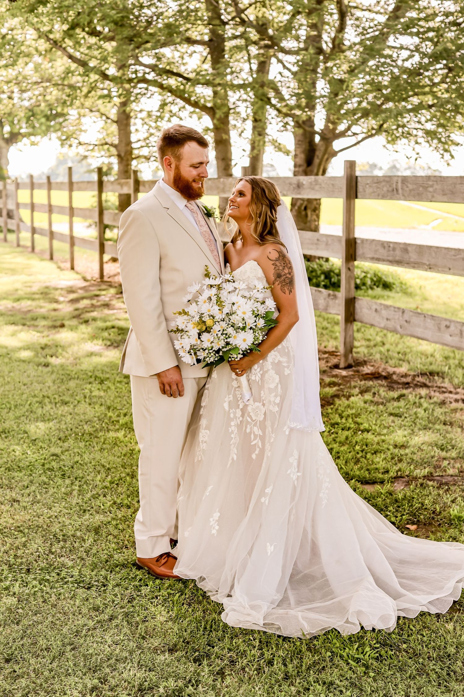A romantic wedding couple poses by a rustic wooden fence in golden sunlight wearing light-colored attire.