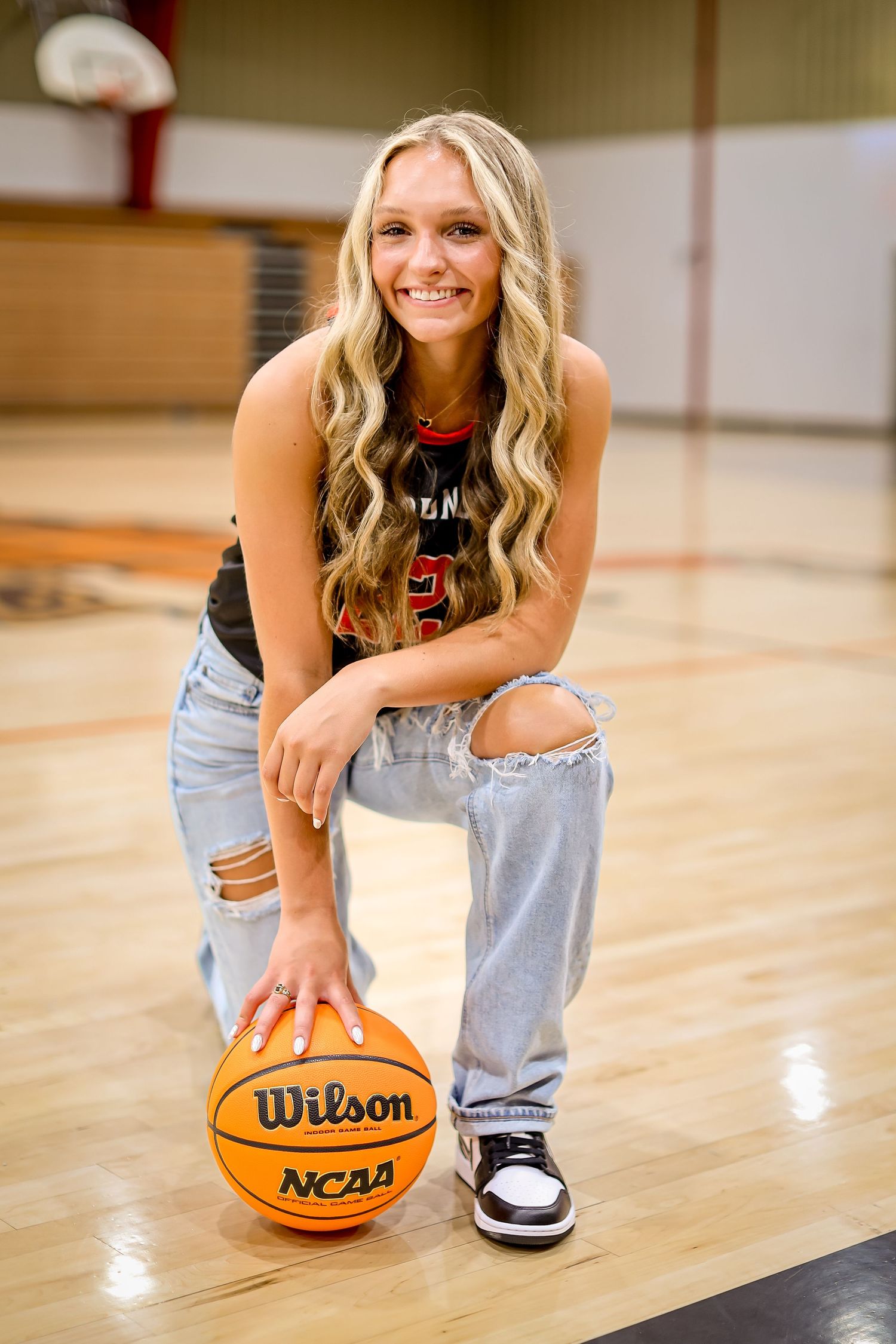 Basketball player posing with Wilson NCAA ball on wooden court in casual ripped jeans.