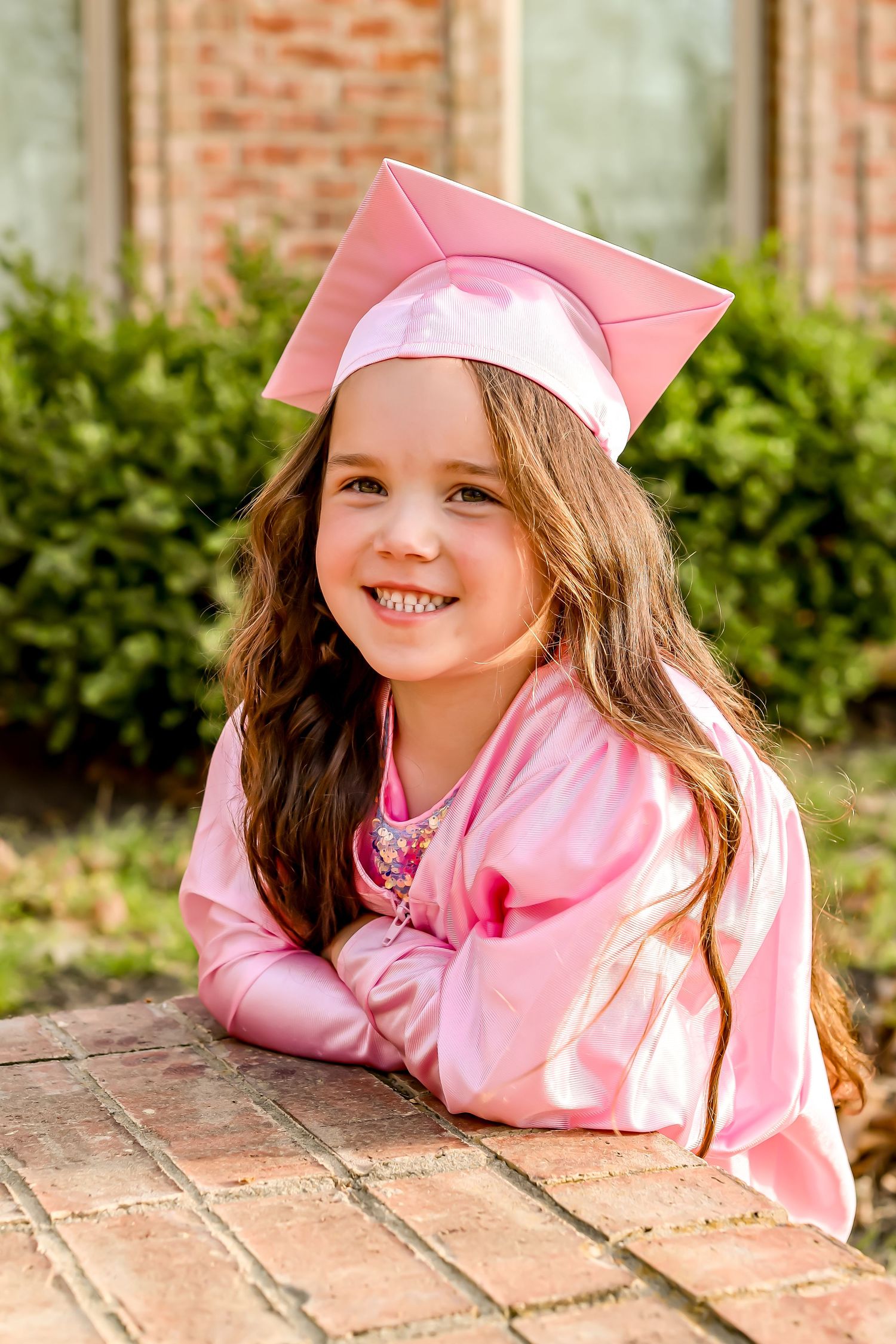 Young graduate in pink cap and gown smiles while leaning on brick ledge.