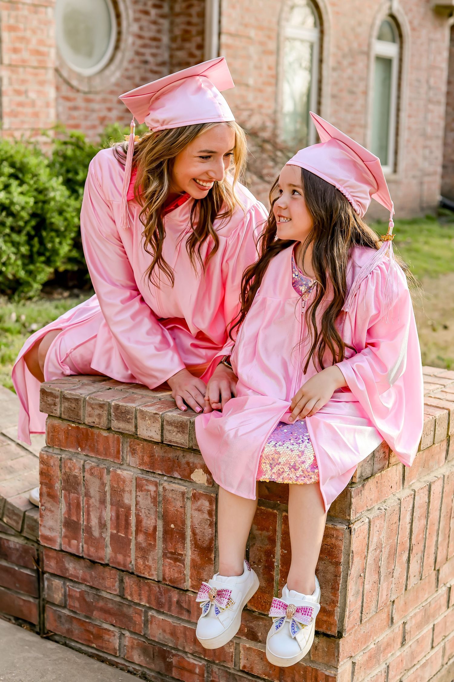 Two graduates in matching pink caps and gowns share happy moment on brick ledge.