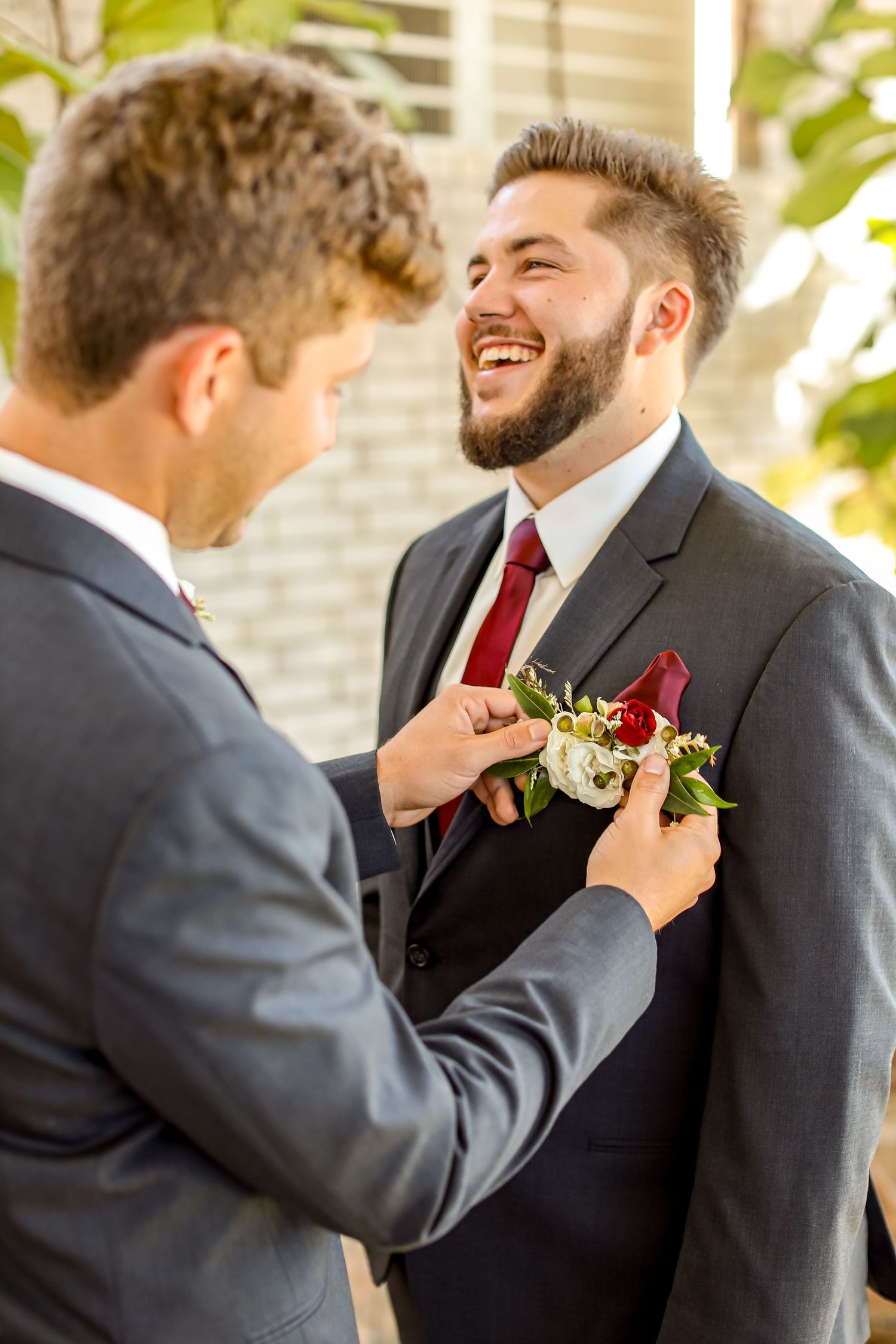 A groomsman helps adjust another's burgundy boutonniere during wedding preparations.