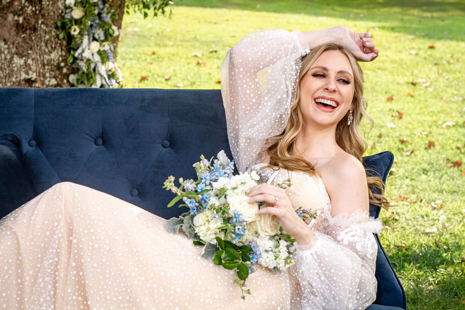 A joyful bride in a sparkly gown relaxes on a navy blue couch outdoors holding a white and blue bouquet.