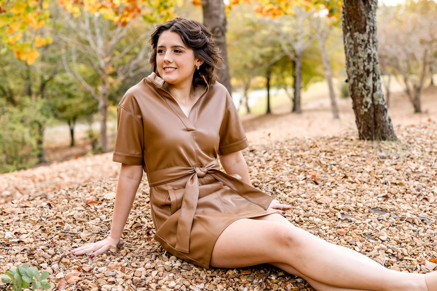 Person in brown dress sitting on ground covered with fall leaves.