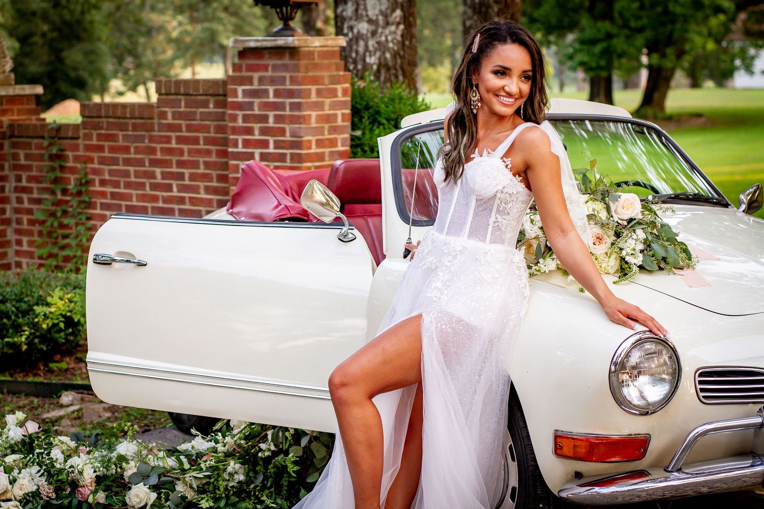A bride in a white dress poses elegantly against a vintage white car on a sunny day.