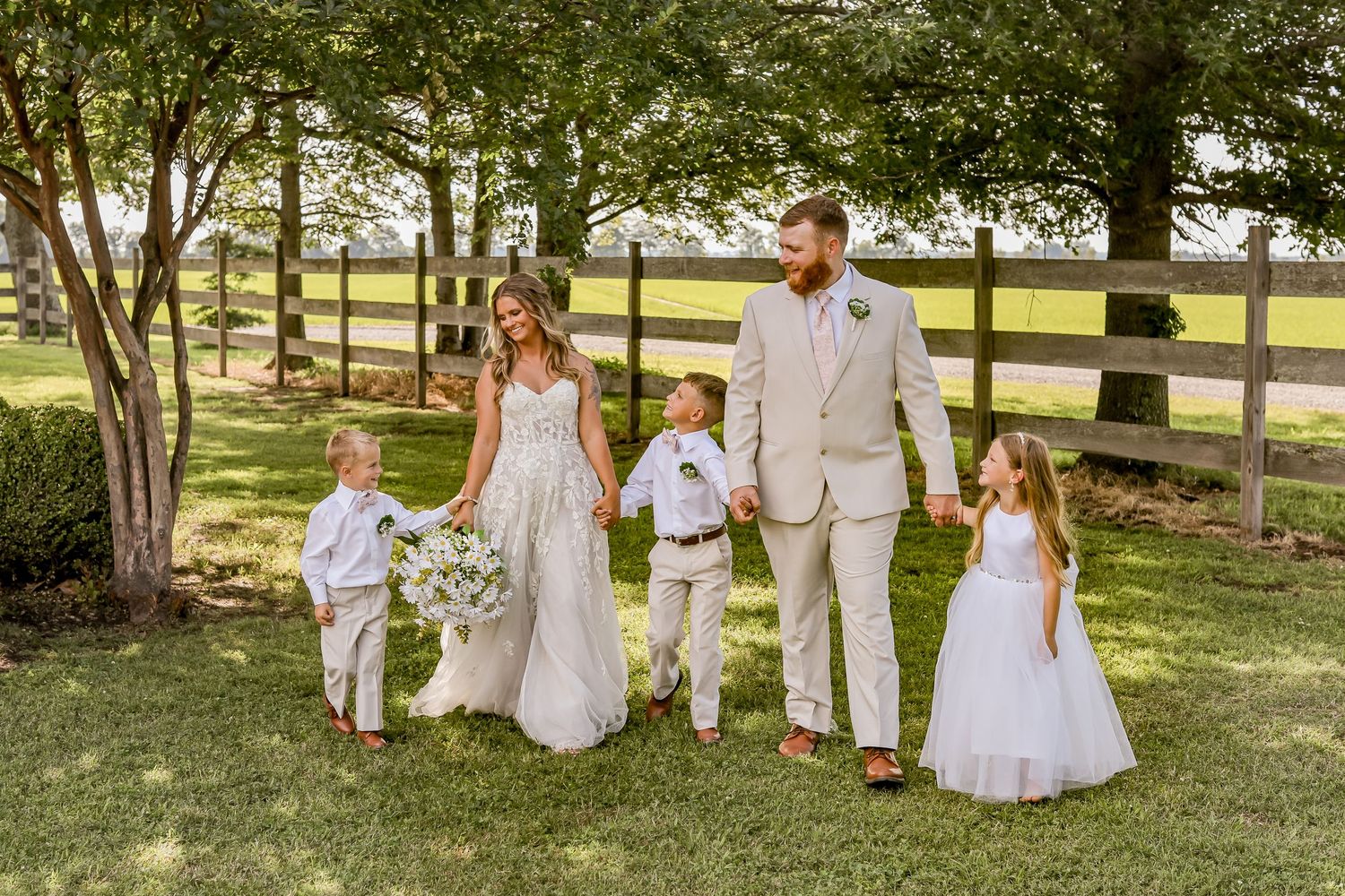 Family in white and beige attire walks together on a sunny lawn with wooden fence.