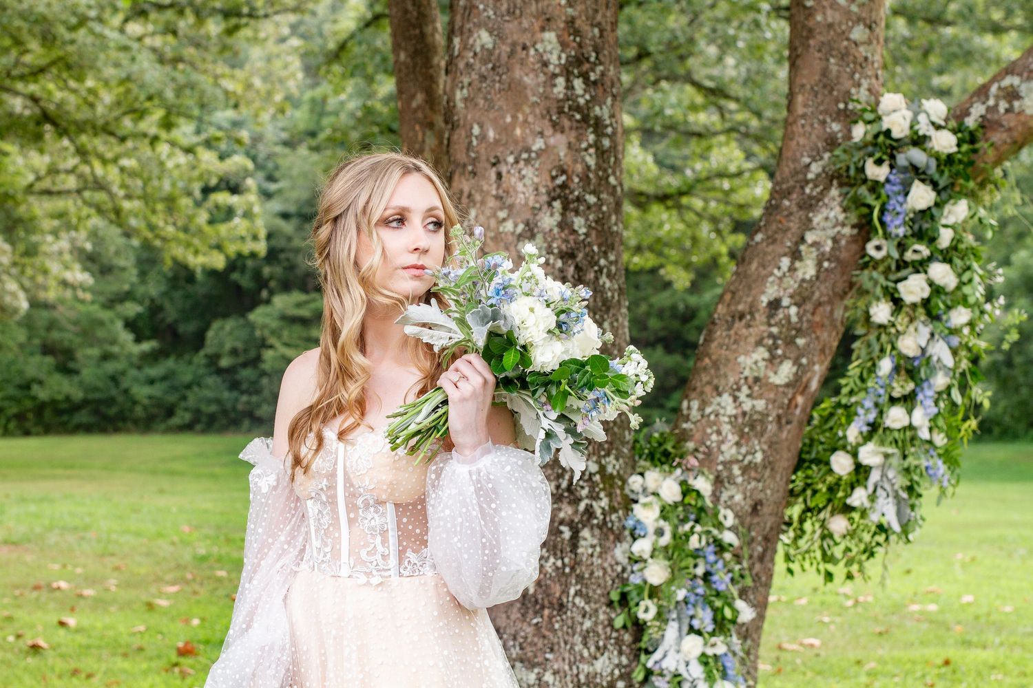 Dreamy garden bridal portrait with white and blue floral bouquet by a tree.