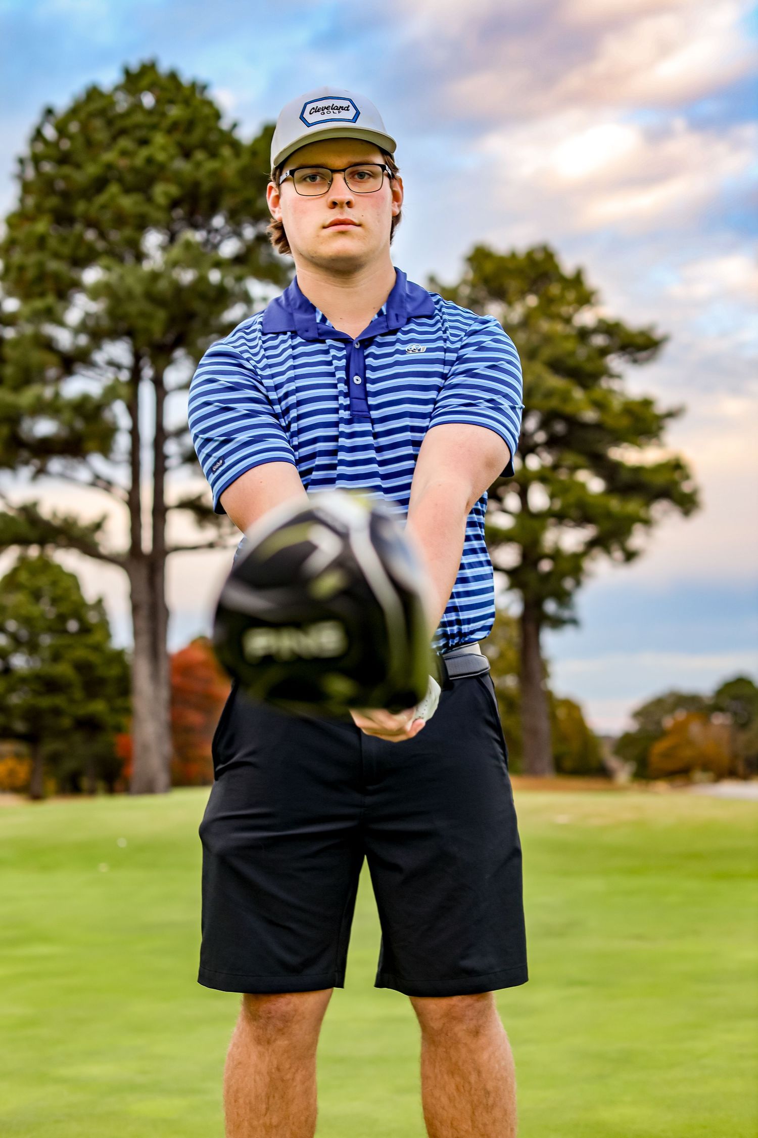 Golfer in striped polo and cap holding club on green course during sunset.