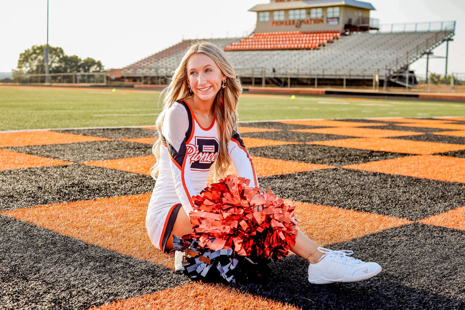 Cheerleader poses with pom poms on sports field with stadium in background.